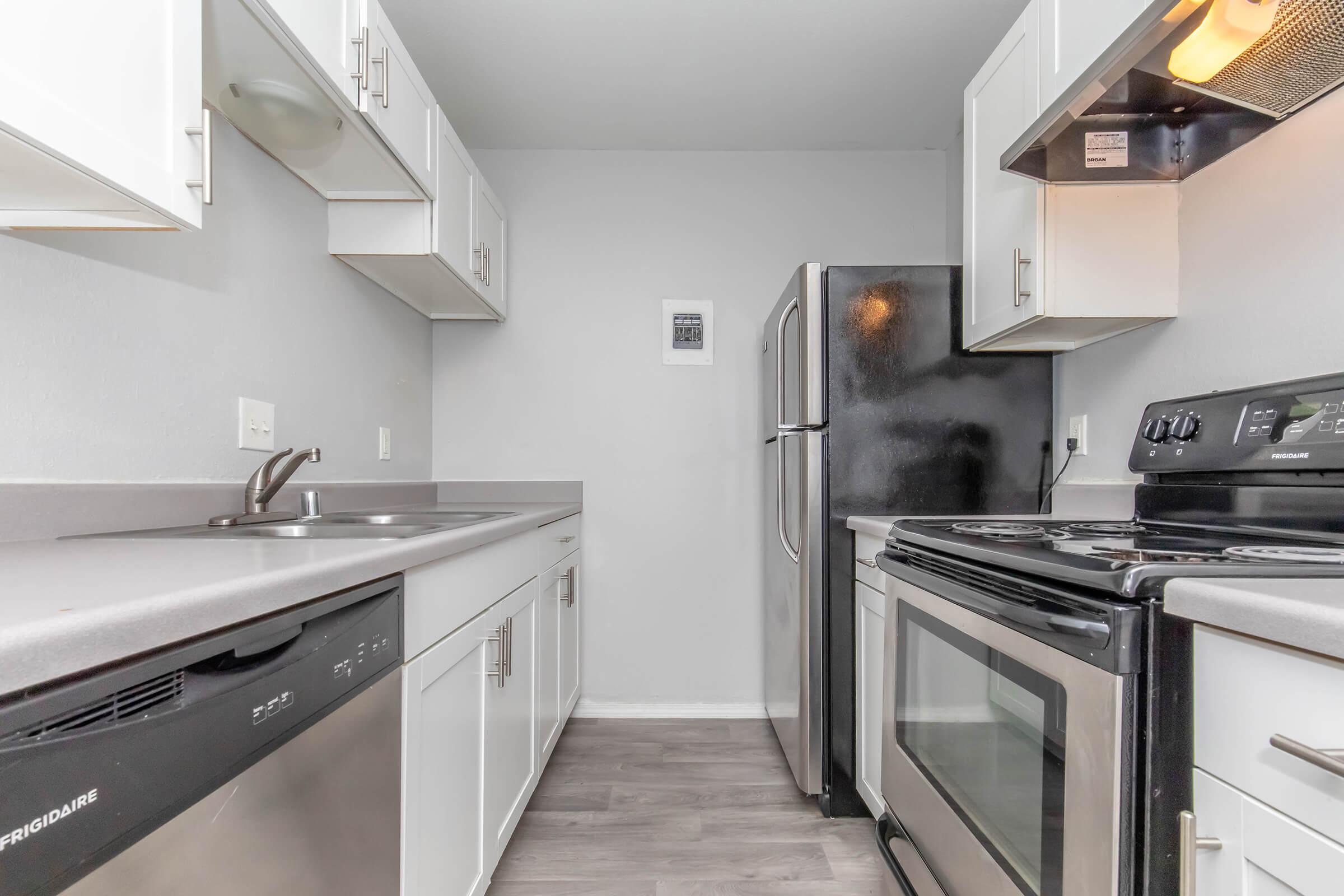 Modern kitchen featuring stainless steel appliances, including an oven, stove, and refrigerator. The cabinetry is white with a gray countertop. The space is well-lit and includes a sink, with a clean, minimalist design. The flooring is a light-colored laminate.