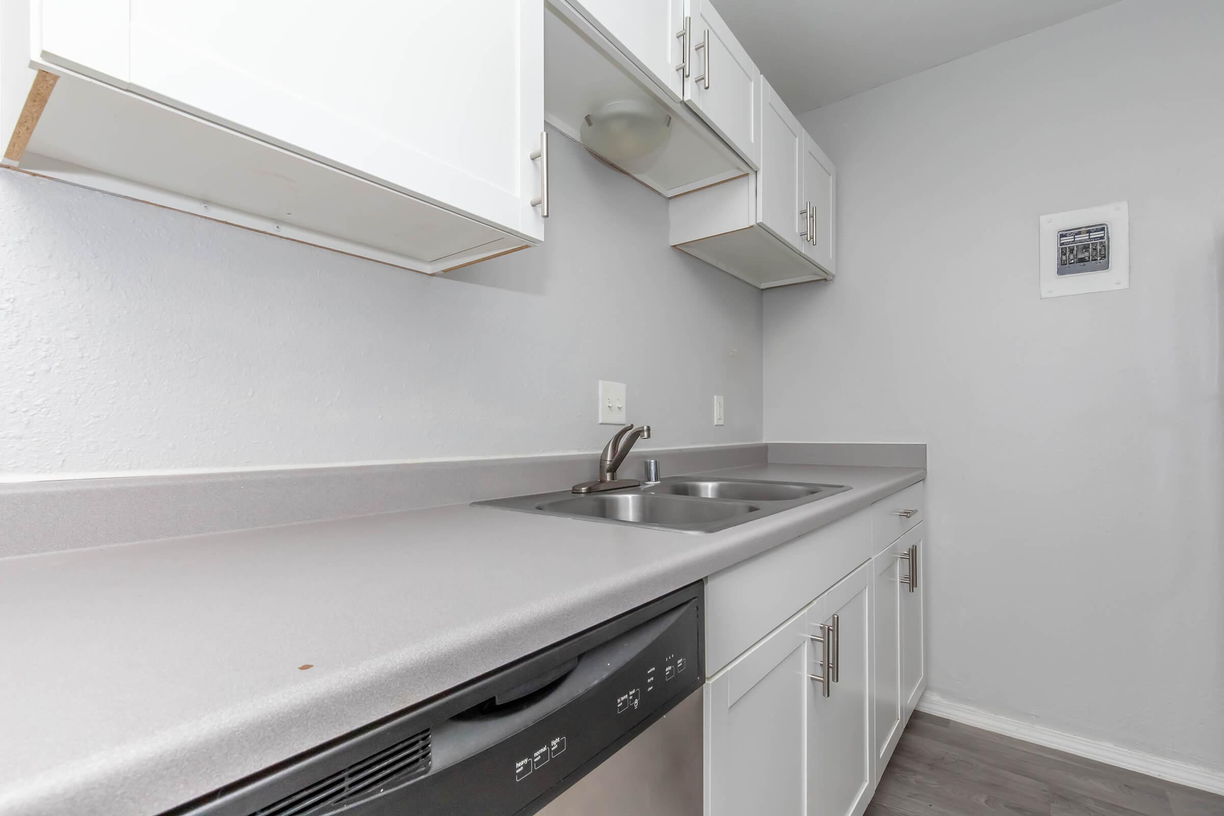 A modern kitchen featuring light gray walls and countertops, with white cabinets above and below. There are dual sinks with a faucet, and a dishwasher at the bottom. The floor has a dark laminate finish, and a framed picture hangs on the wall. The overall aesthetic is clean and minimalistic.