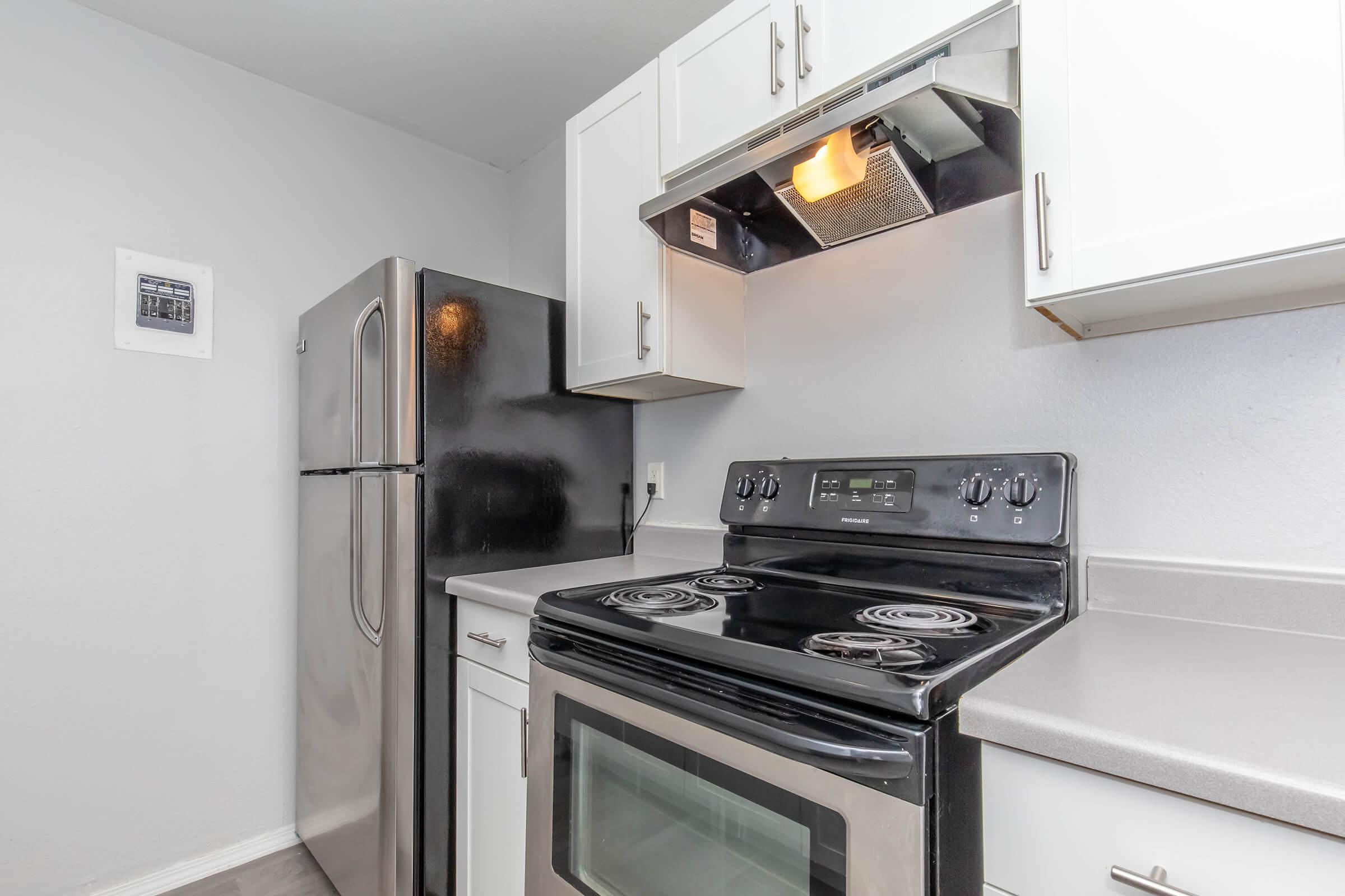 A modern kitchen featuring stainless steel appliances including a refrigerator and an oven with a cooktop. The walls are painted light gray, and the cabinets above the countertop are white. A range hood is installed above the stove, providing ventilation. Overall, the space appears clean and well-lit.