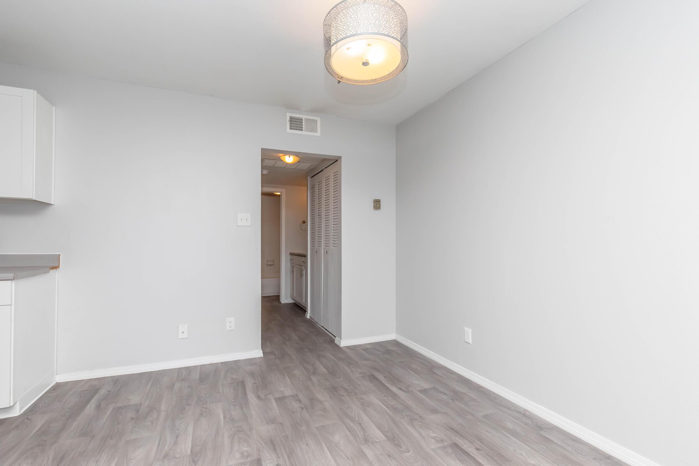 A spacious, modern room with light gray walls and flooring. It features a simple ceiling light fixture and an open doorway leading to a hallway. The area is uncluttered, showcasing potential for furniture arrangement.
