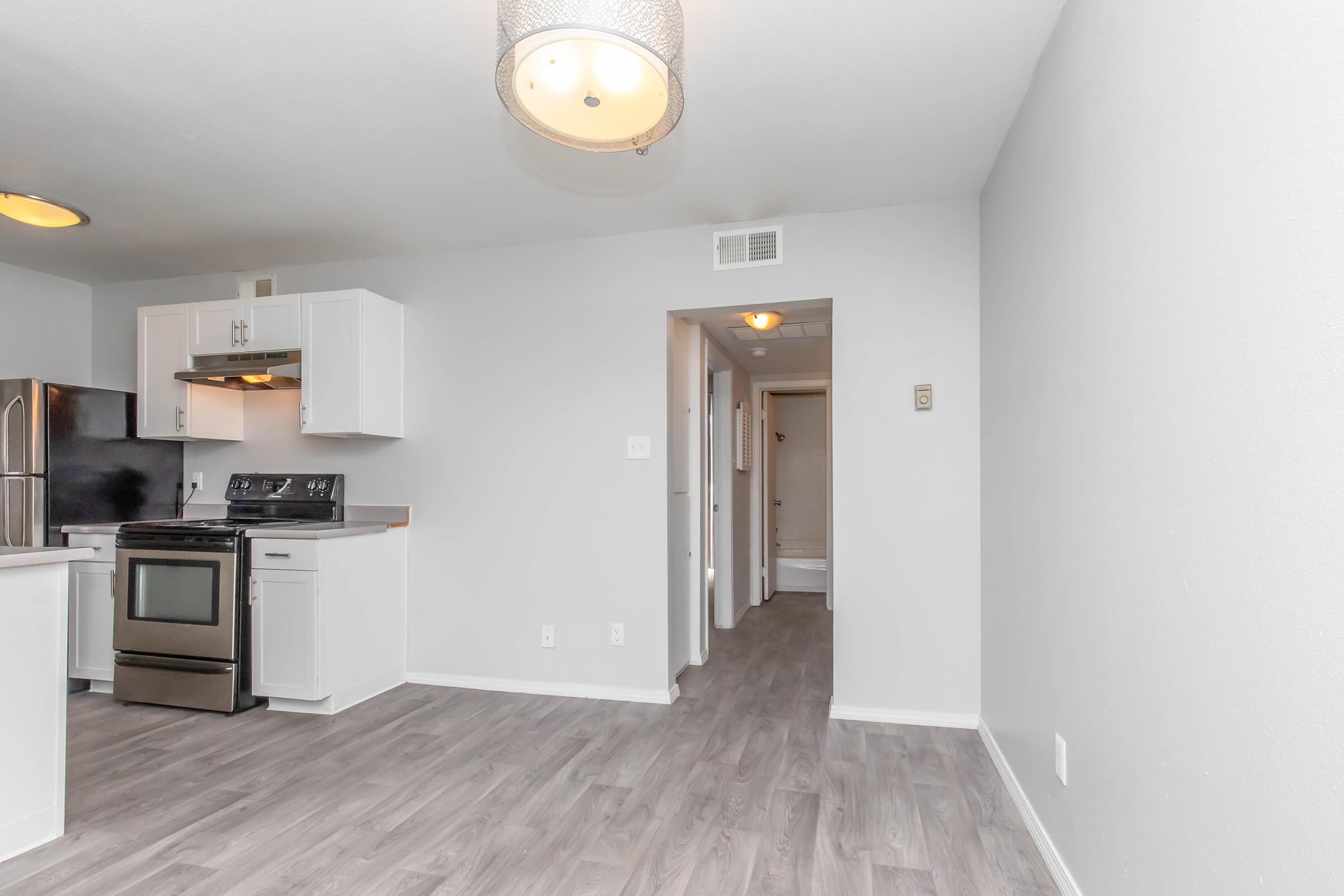 A modern kitchen with white cabinets, a stainless steel stove, and a refrigerator. Light-colored walls and laminate flooring create a spacious feel. A doorway leads to a hallway, and a ceiling light fixture adds illumination to the room.