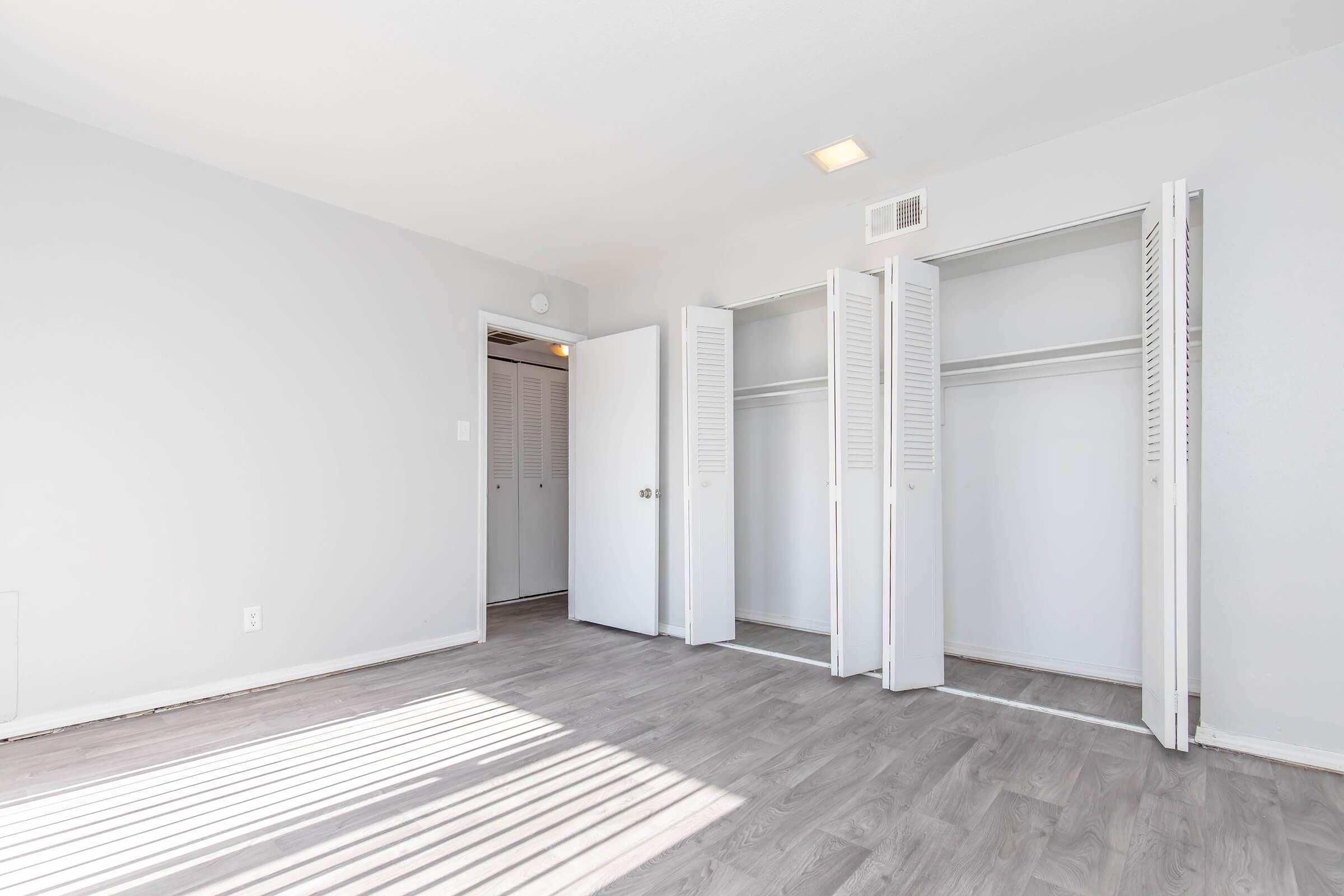 A bright, empty room with light gray walls and light wood flooring. Two open closet doors on the right reveal empty spaces. A door to the left leads to another area. Sunlight casts shadows on the floor from the windows.