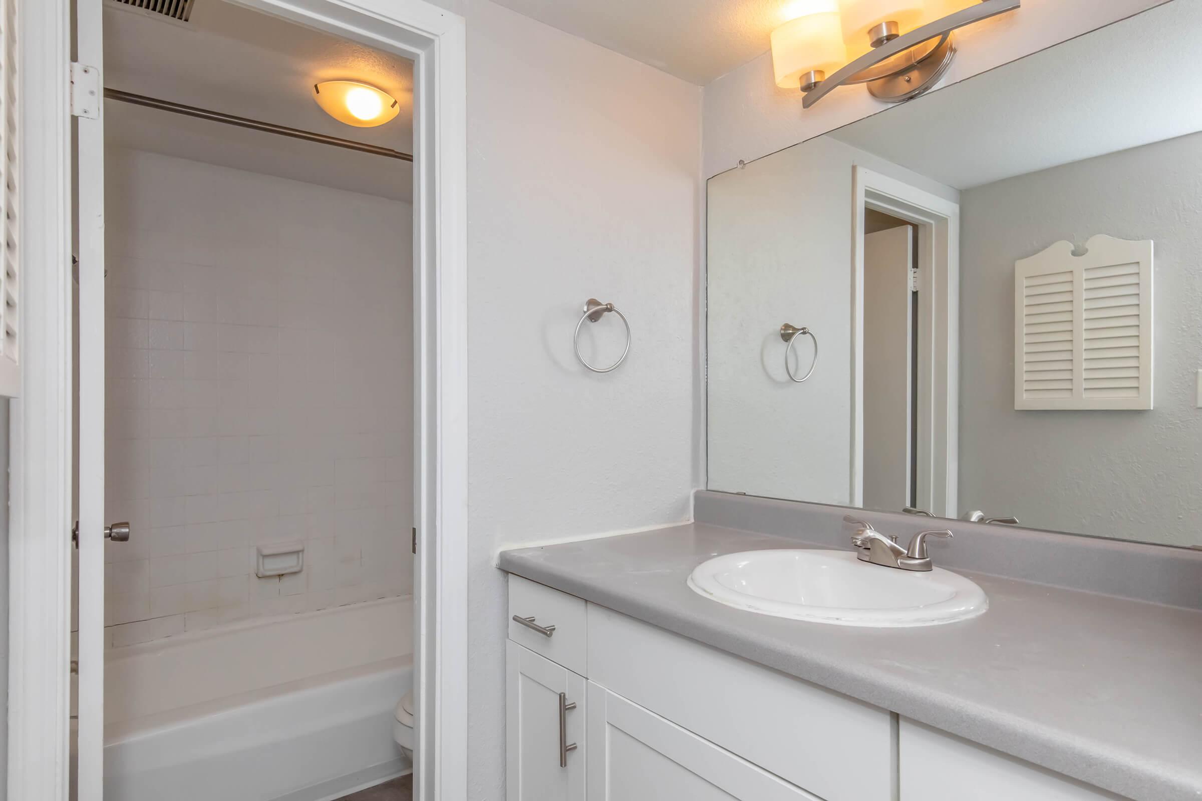 A clean and modern bathroom featuring a white vanity with a sink, a large mirror above, and a light fixture. To the left, a doorway leads to a bathtub area with tile walls, while the right side includes a towel rack. The walls are painted a soft color, contributing to a bright and airy atmosphere.