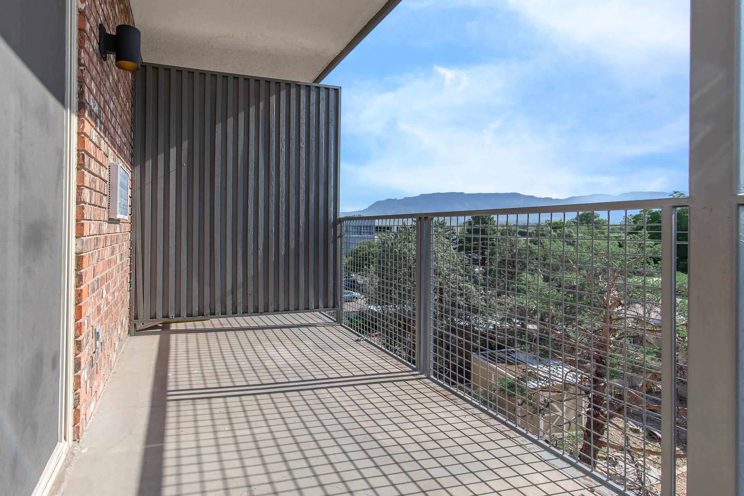 A modern balcony with a metal railing and a sliding privacy screen, featuring a view of distant mountains and lush greenery. The floor is concrete, and the wall shows exposed brick. Soft natural light illuminates the space, creating a serene atmosphere.