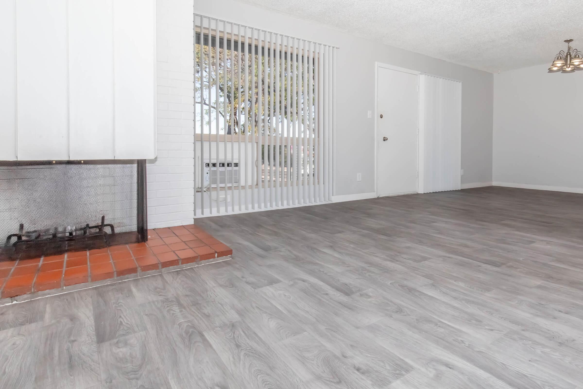 Interior of a living room featuring gray laminate flooring, a white brick fireplace with a red brick hearth, large windows with vertical blinds, and a door leading to another room. The walls are painted light gray, contributing to a bright and open atmosphere.