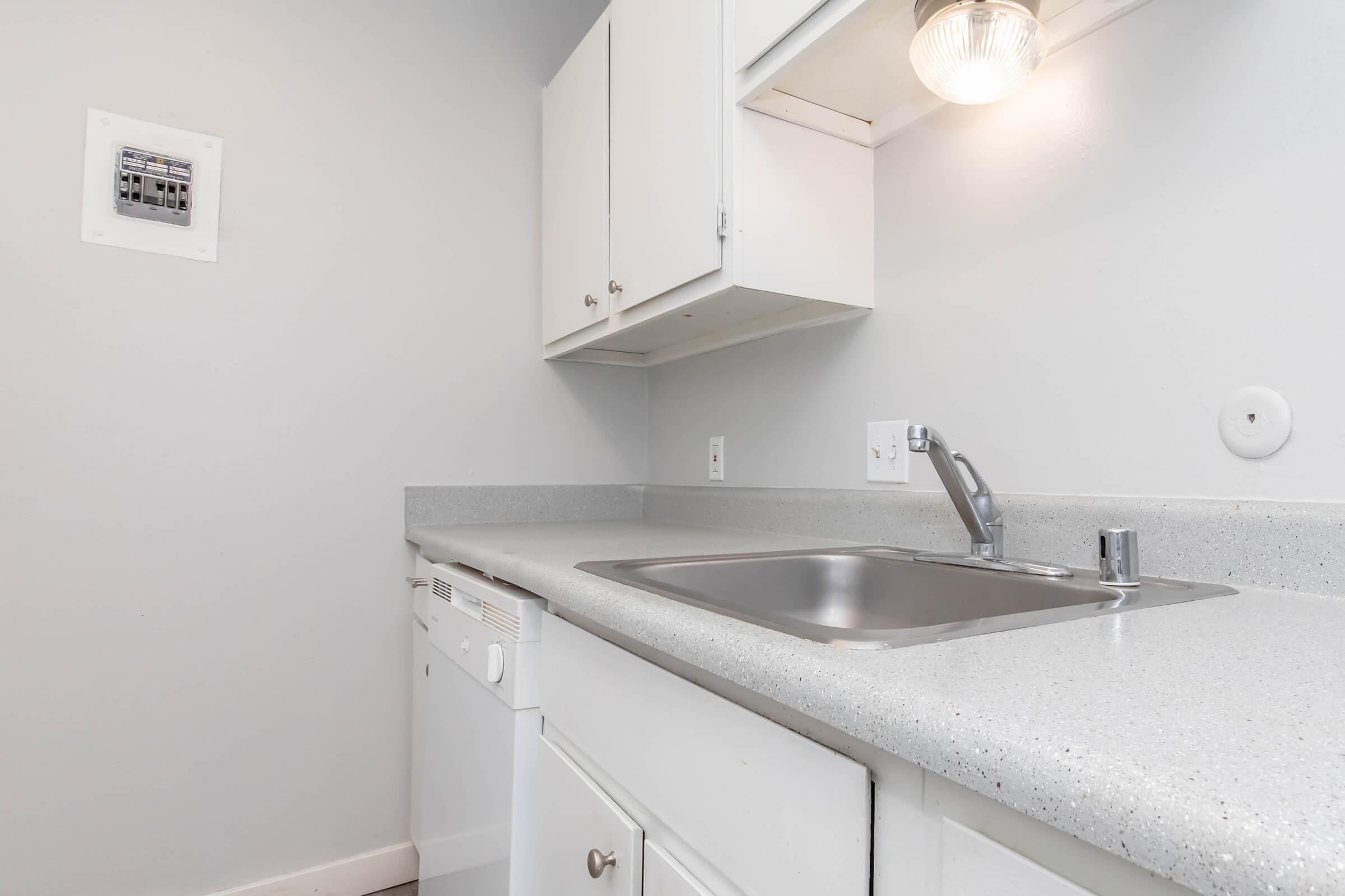 A small, modern kitchen featuring a stainless steel sink, a white countertop, and a built-in dishwasher. The cabinets above and below are white, and there is a light fixture above the sink providing illumination. The wall is painted in a light gray color, creating a clean, airy atmosphere.