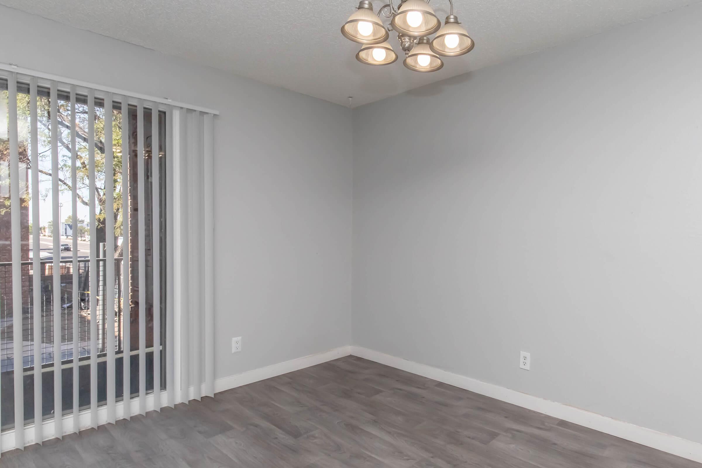 A well-lit, empty room featuring light gray walls and a wooden floor. There is a ceiling light fixture with multiple bulbs and a sliding glass door with vertical blinds, providing access to an outdoor area. The room appears spacious and ready for furniture.
