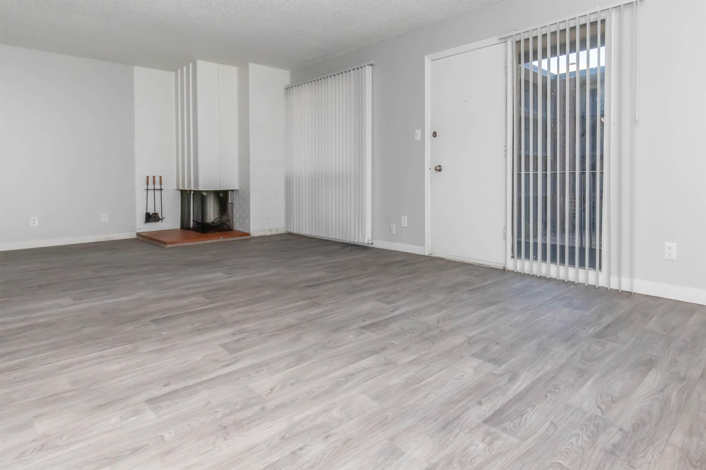 A spacious, empty living room featuring light gray walls, a hardwood floor, and a large window with vertical blinds. There is a fireplace with a white mantel on one side and a door leading outside on the other. The room is well-lit, creating an airy atmosphere.