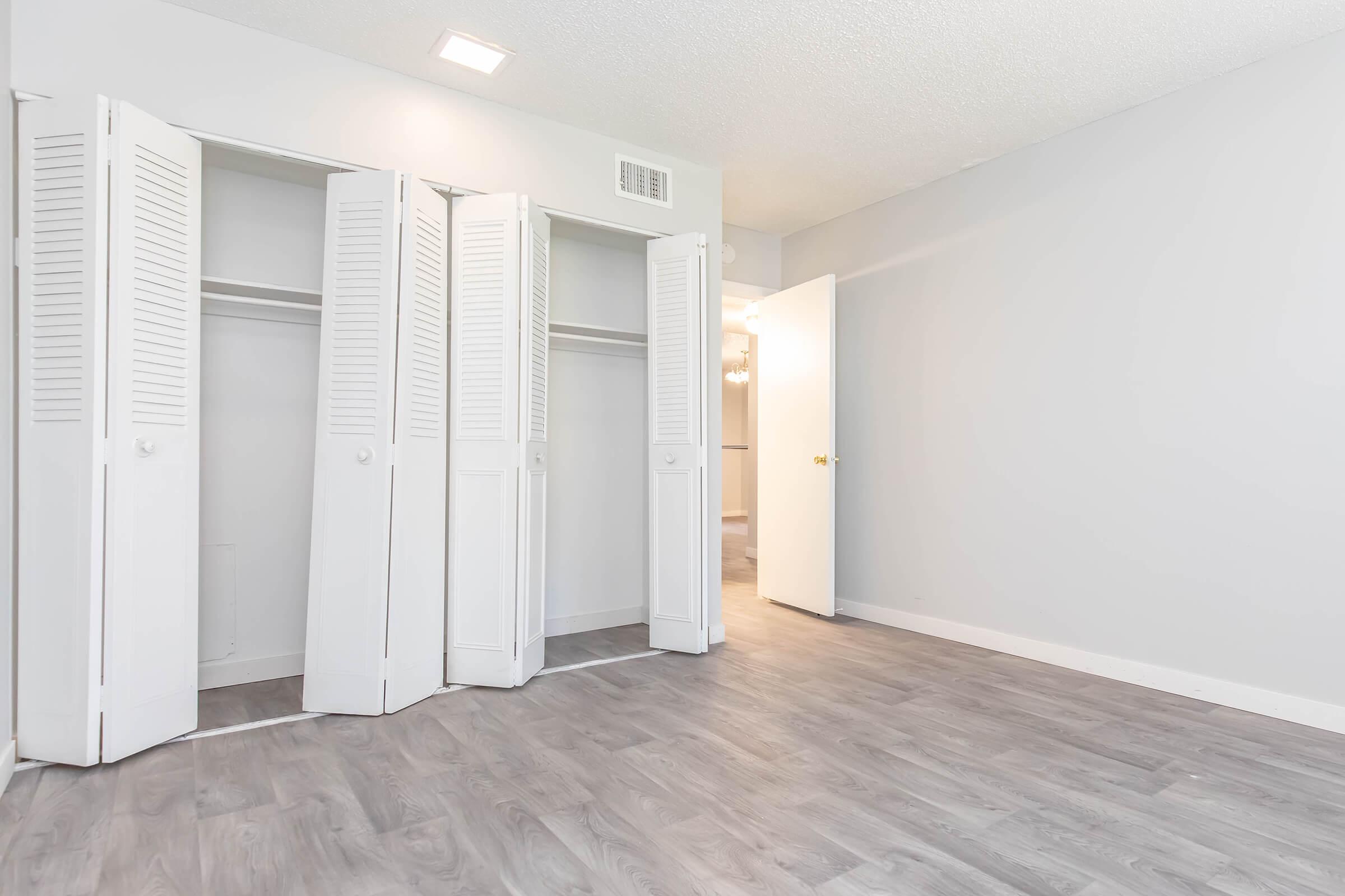 A room featuring light gray walls and wooden flooring. There are two sets of white louvered closet doors along one wall, partially open, revealing empty storage space inside. A door on the right leads to another area, with soft lighting visible in the background. The overall atmosphere is bright and minimalist.