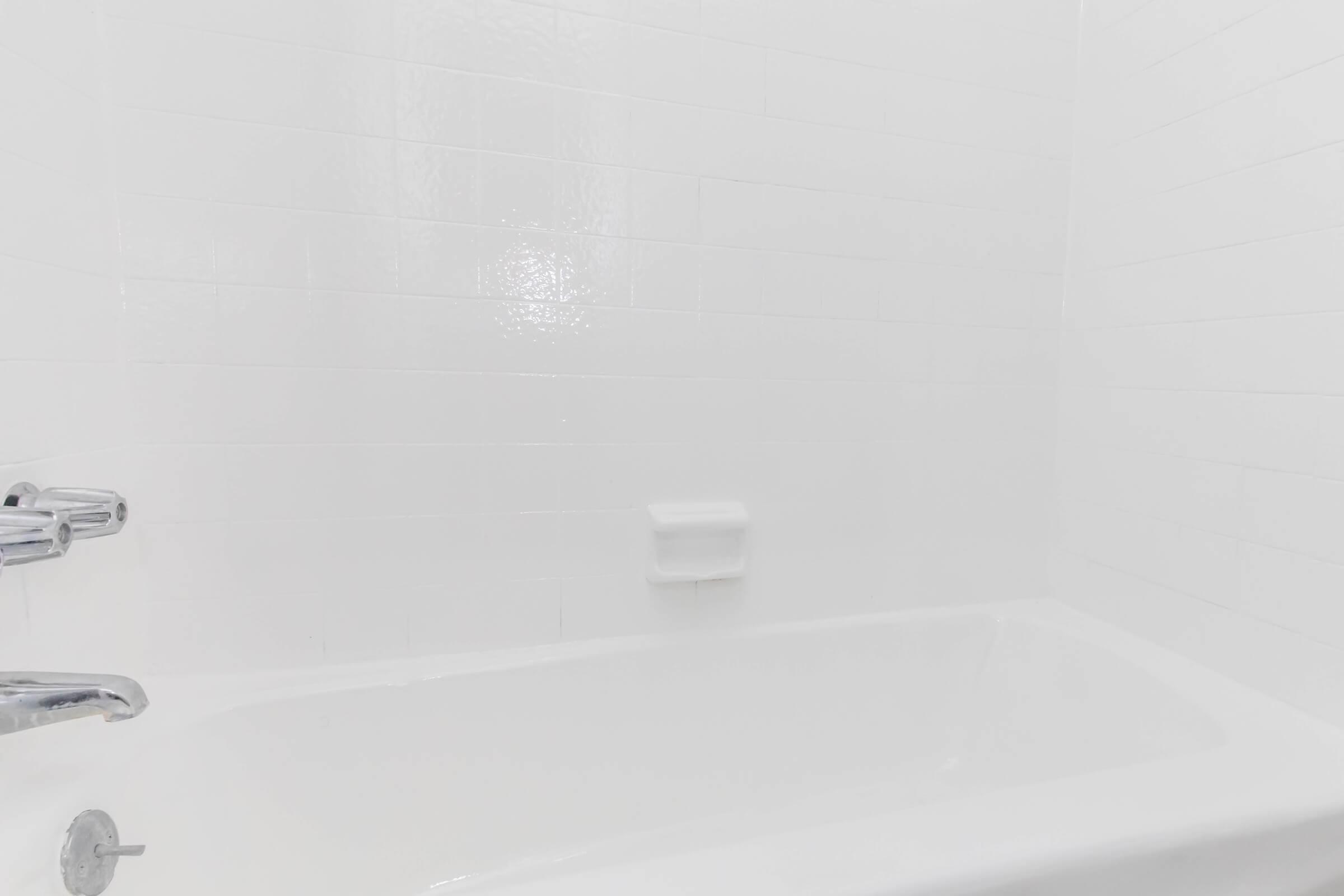 A clean, modern bathroom featuring a white bathtub with a chrome faucet. The walls are tiled in a glossy white finish, creating a bright and minimalist aesthetic. A small soap holder is visible on the wall, contributing to the simple and uncluttered design.