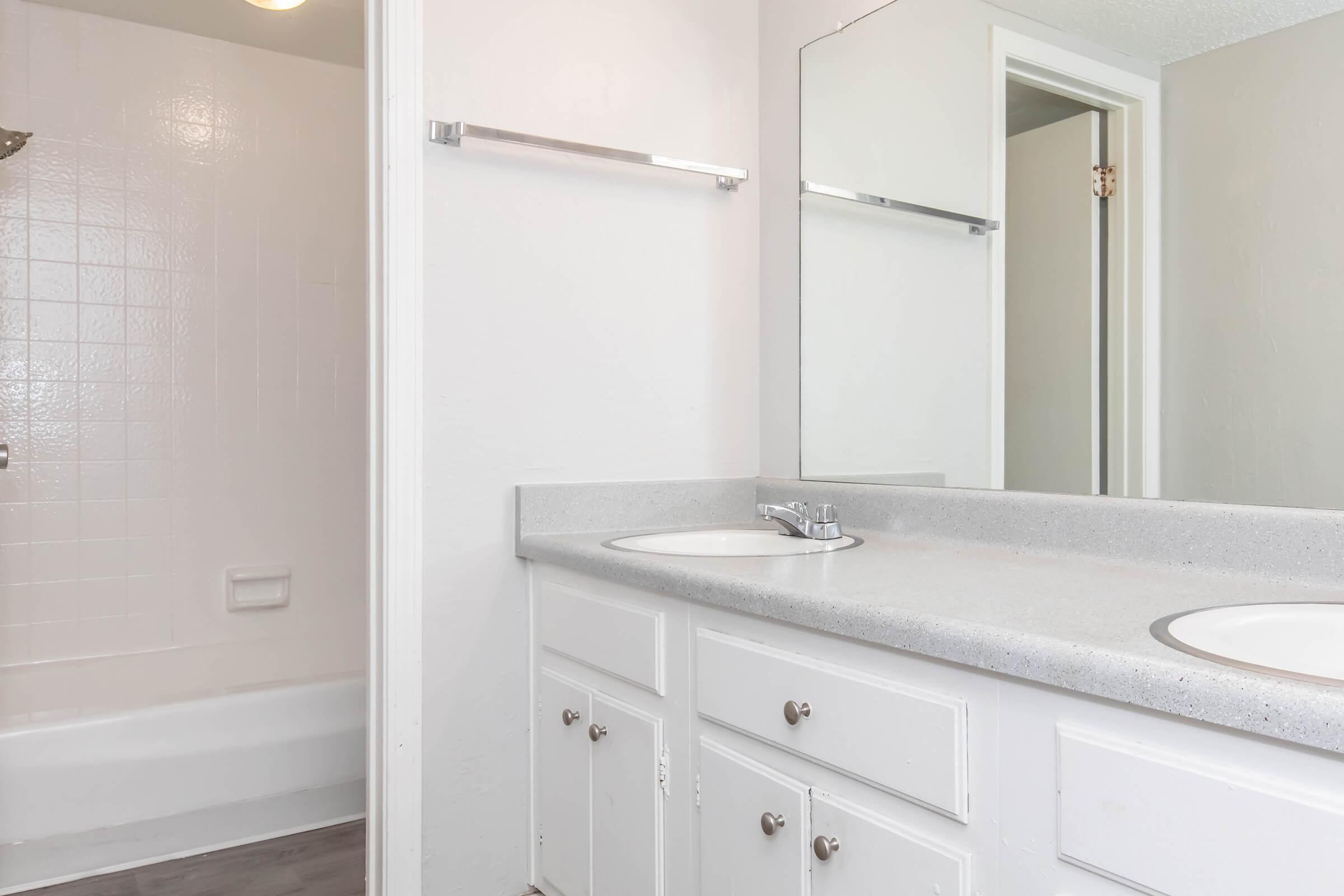 A clean and modern bathroom featuring a light-colored countertop with dual sinks and silver fixtures. The walls are painted in a neutral tone, and there's a bathtub with a tiled wall visible in the background. A mirror hangs above the sinks, reflecting the bright, spacious room.