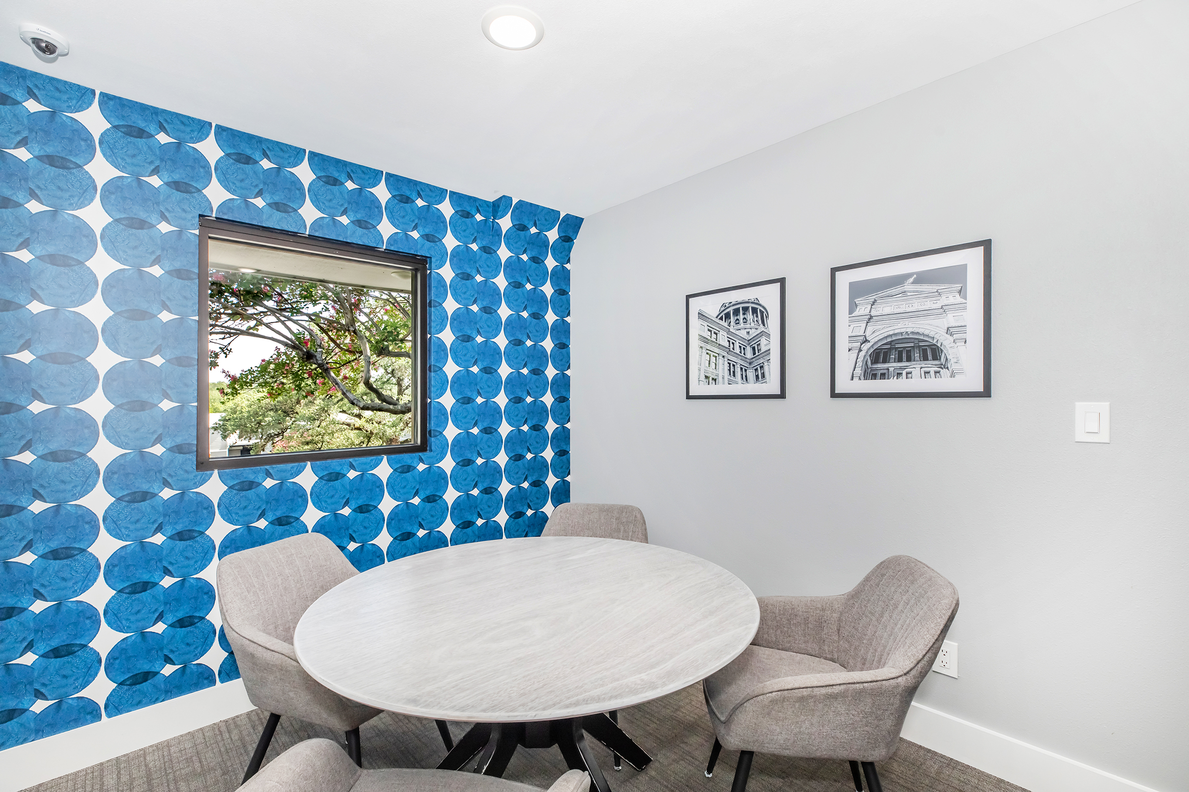 A modern meeting room featuring a round table surrounded by four gray chairs. The walls are decorated with a vibrant blue patterned design. A window with a view of trees brings natural light into the space. Two framed black-and-white photographs adorn the wall.
