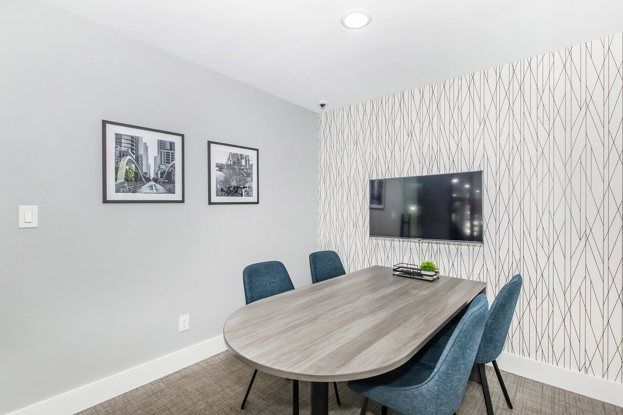 A modern conference room featuring a round wooden table surrounded by four blue upholstered chairs. On the walls, there are two black-and-white framed photographs of urban scenes, and a flat-screen TV is mounted on one wall. The room has a minimalist design with a neutral color palette.