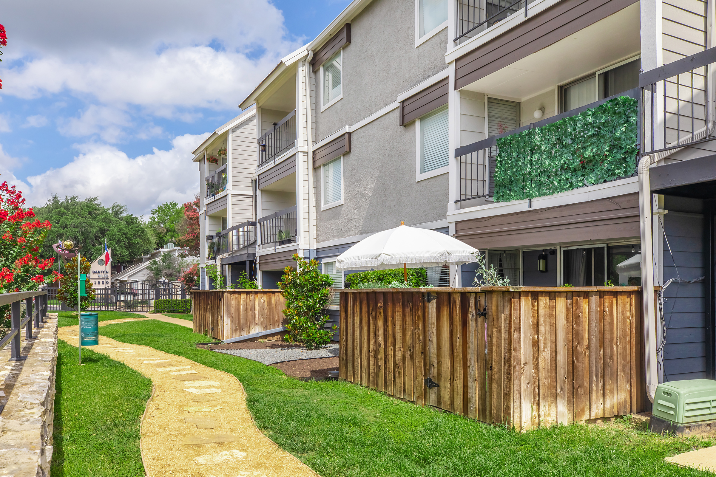 View of a multi-unit apartment building with balconies. The pathway is lined with grass and leads to a wooden fence enclosing small outdoor spaces. Flowering plants and a white umbrella on a patio add to the scenery. Bright blue skies with scattered clouds enhance the setting.