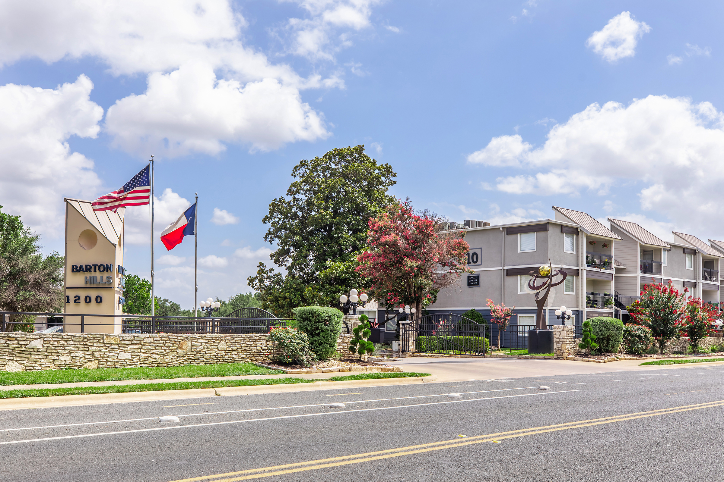 Sign for Barton Hills with the address "1200" and American and Texas flags flying nearby. The entrance features well-maintained landscaping with trees and flowering plants. In the background, there is a multi-story building with balconies, under a partly cloudy blue sky.