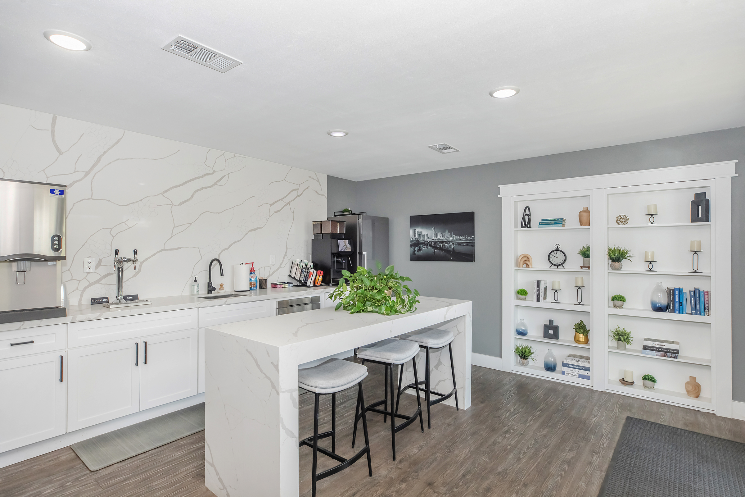 Modern kitchen featuring a large white marble island with three gray bar stools, sleek cabinetry, and a minimalist design. There's a decorative plant on the island, a coffee maker, and a shelf unit displaying books and decor items against a neutral wall backdrop. Bright lighting enhances the spacious feel.
