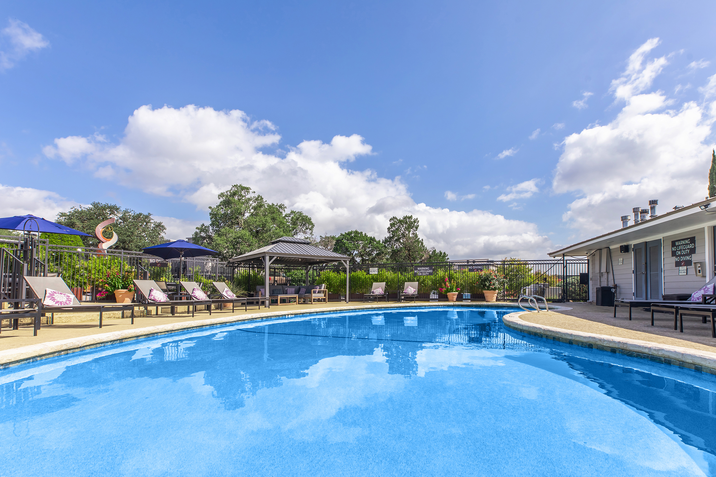 A bright, sunny day at a swimming pool surrounded by lounge chairs and umbrellas. The clear blue water reflects the sky and nearby trees. In the background, there are pavilions and a building with a sign, all under a partly cloudy sky.