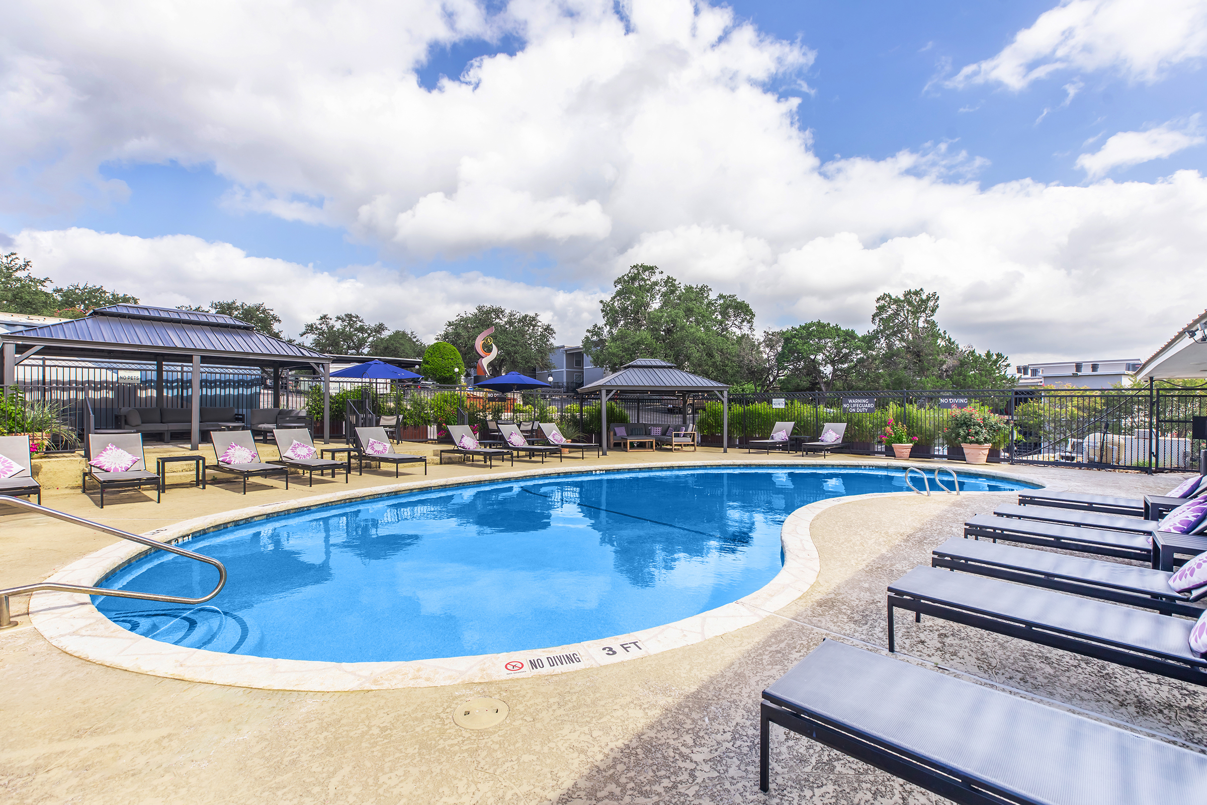 A serene outdoor pool area featuring a large, clear blue pool surrounded by lounge chairs. There are decorative plants and trees in the background, along with shaded seating areas under gazebos. The scene is set against a partly cloudy sky, creating a relaxing atmosphere.