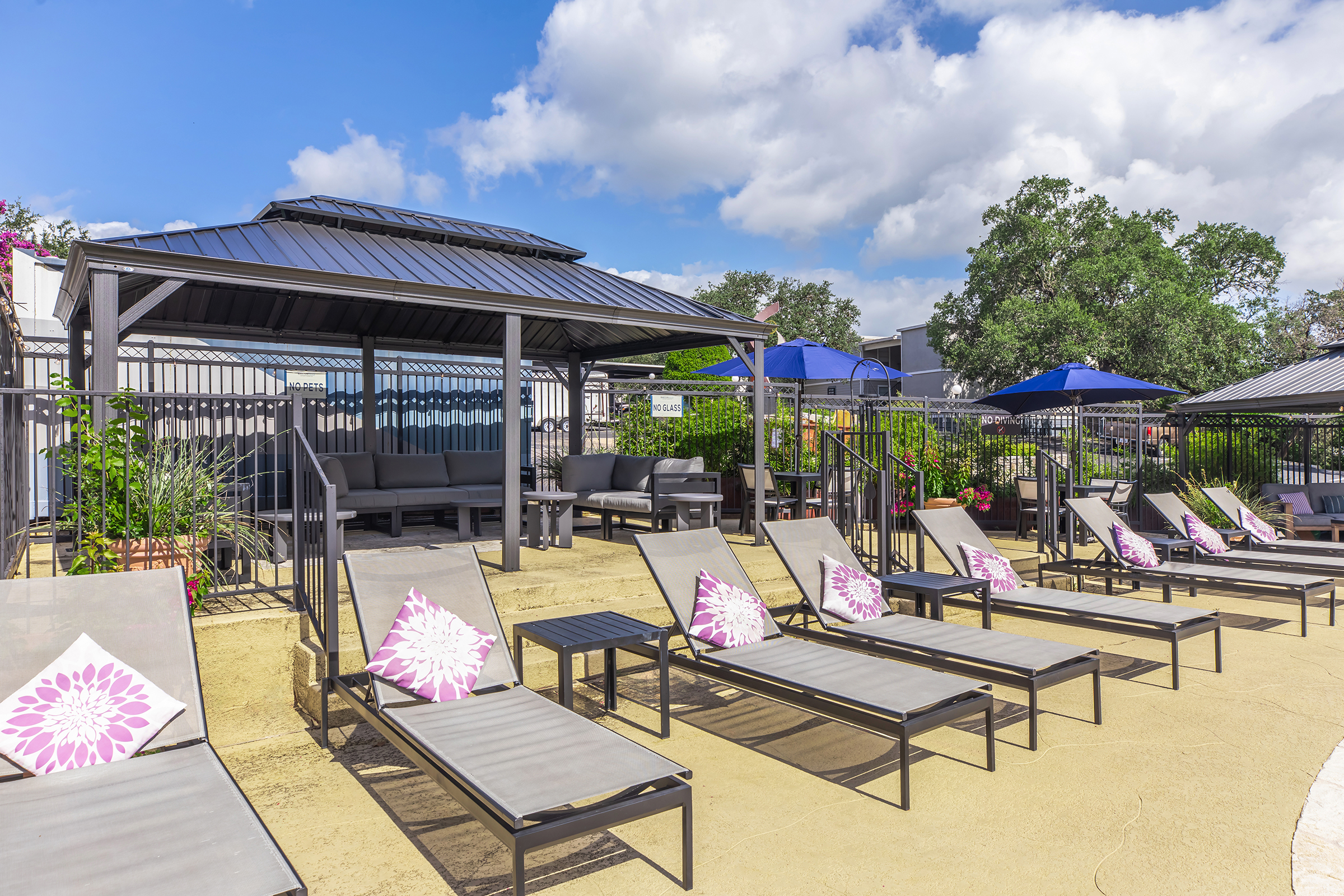 A sunny pool area featuring several lounge chairs with purple floral cushions, private cabanas, and blue umbrellas. Lush greenery surrounds the space, and a clear blue sky with fluffy clouds adds to the relaxing atmosphere.