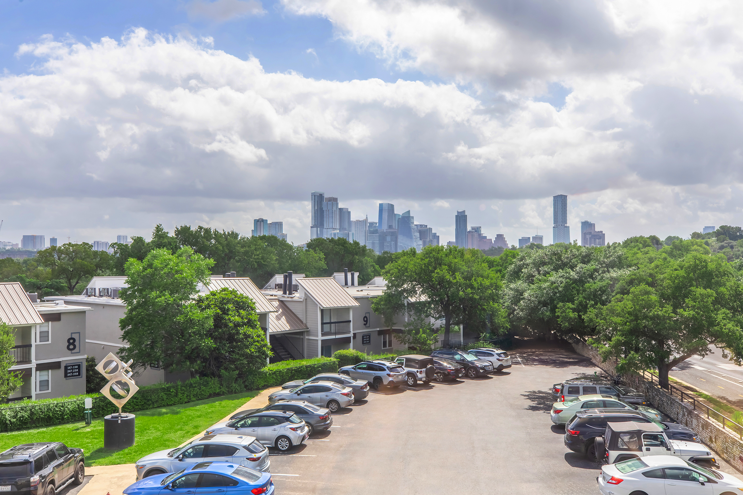 A view of a parking lot with various cars parked, set against a backdrop of a city skyline featuring tall buildings. The scene is partly cloudy with greenery in the foreground, including trees. The image conveys a mix of urban and natural elements.