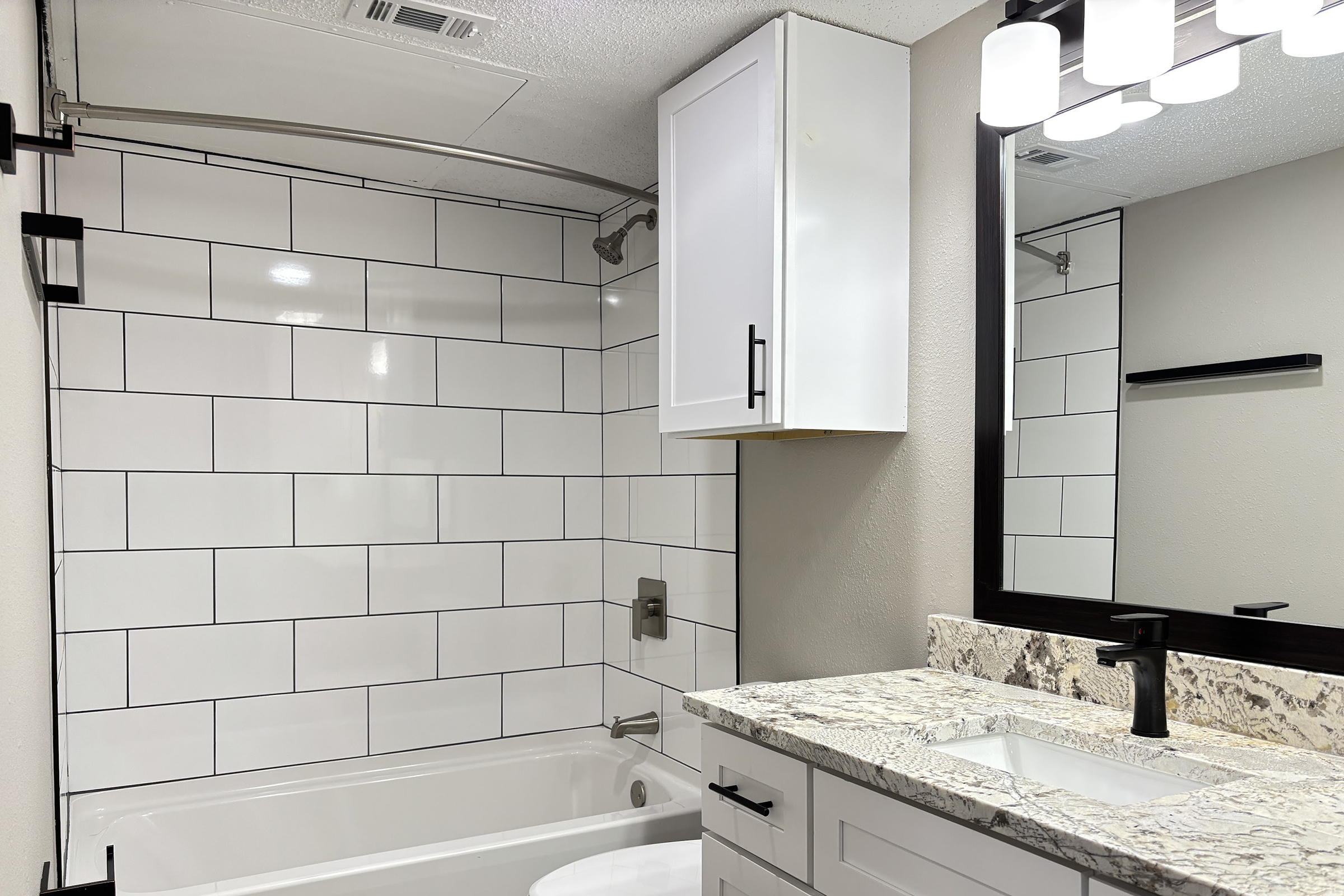 Modern bathroom featuring white tiled walls, a bathtub, and a sleek countertop with a sink. The mirror above the sink is framed in black, and there are contemporary light fixtures above. A white cabinet is mounted on the wall. The overall design is clean and minimalist.