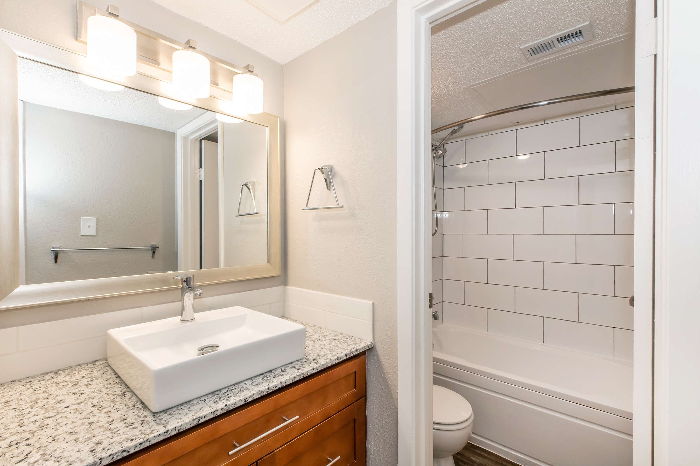 A modern bathroom featuring a light grey color palette. It includes a large mirror above a stylish white sink with a granite countertop, wooden cabinetry, and chrome fixtures. A bathtub with white tiled walls is visible through an open doorway, and there are wall-mounted towel fixtures.