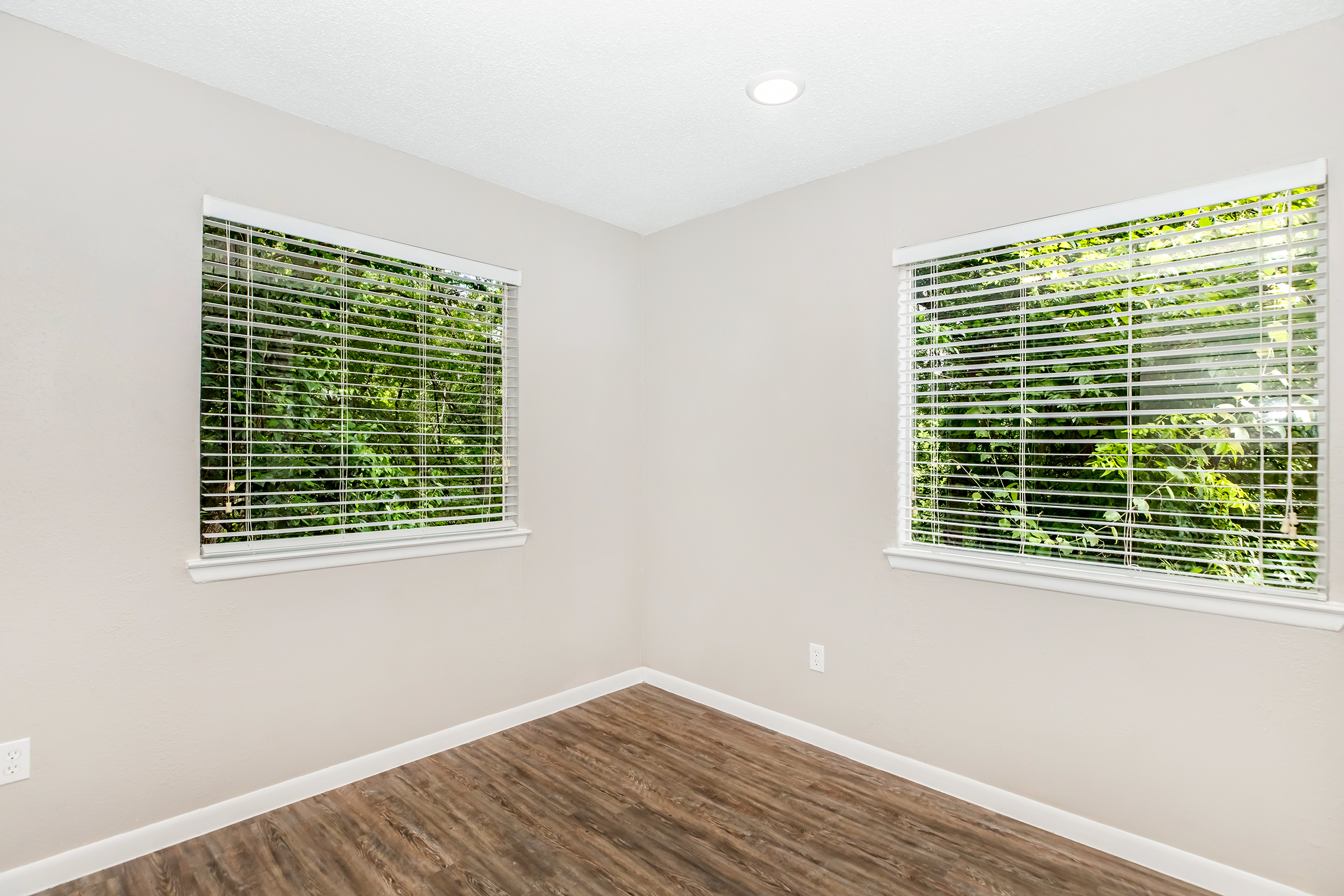 A light-colored room with two windows featuring white blinds, showcasing a view of green foliage outside. The floor is a warm wooden tone, and the walls are painted a neutral shade. The room is empty and well-lit, creating a spacious feel.