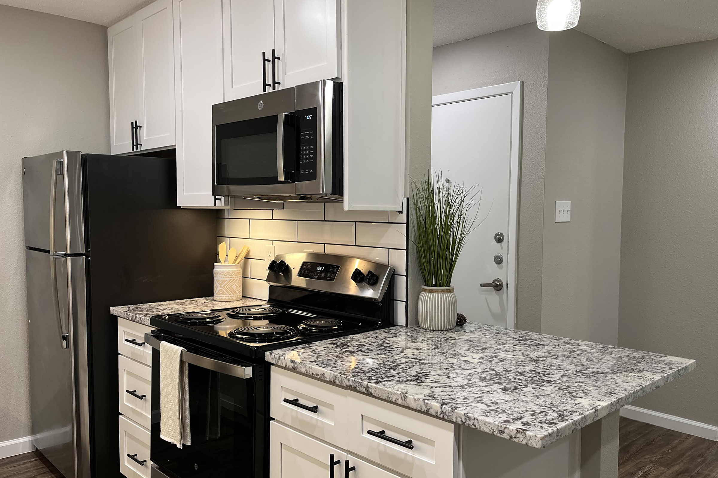Modern kitchen featuring white cabinets, stainless steel appliances, and a granite countertop. A black refrigerator is on the left, and an electric stovetop with an oven is on the right. There is a small potted plant on the countertop, and the wall is tiled with white subway tiles. A door is visible in the background.