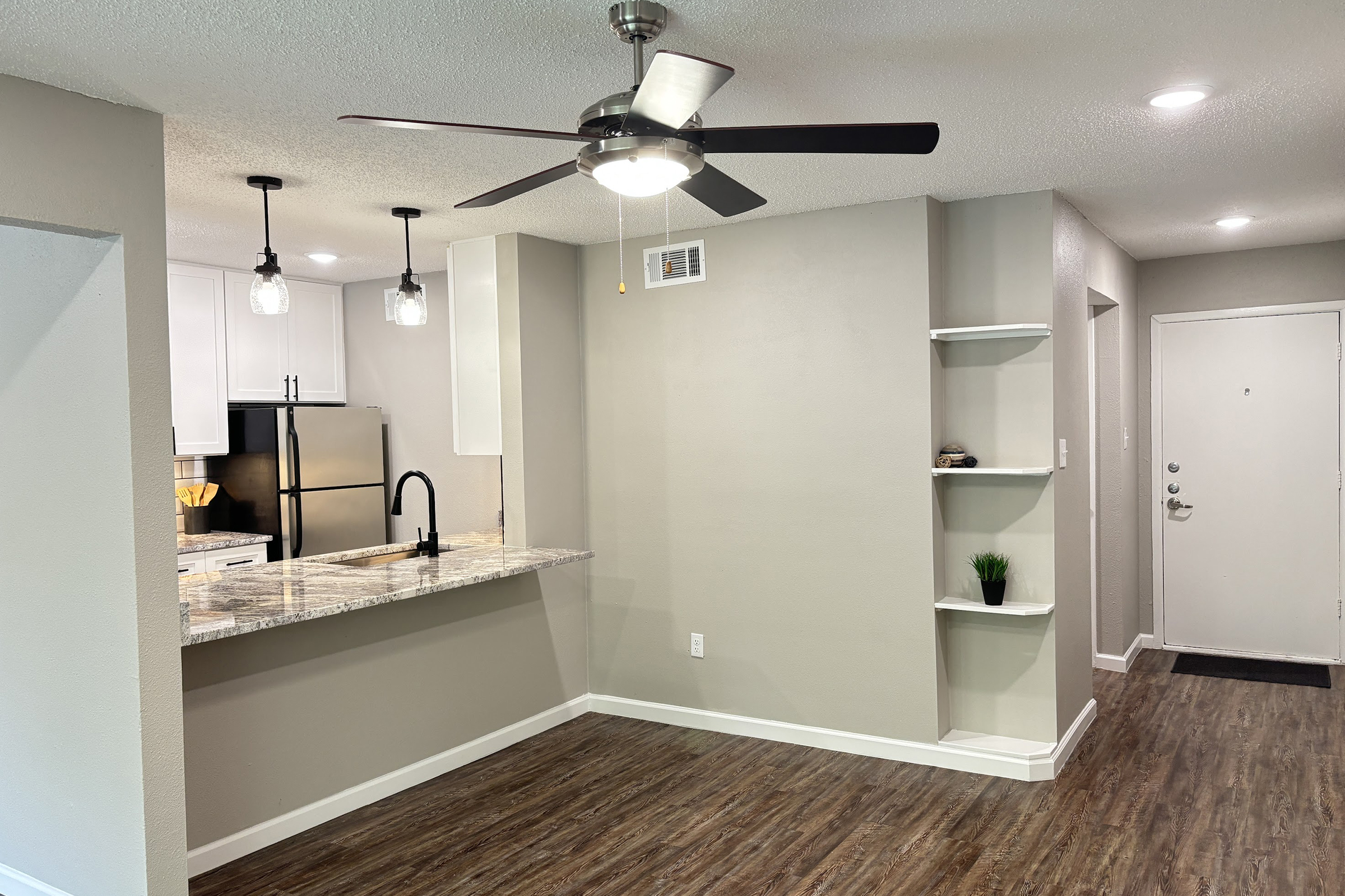 Open concept living space featuring a modern kitchen with a granite countertop and stainless steel appliances. There is a ceiling fan for ventilation, stylish pendant lighting, and a decorative shelf along one wall. The entrance door is visible in the background, along with laminate flooring throughout the area.