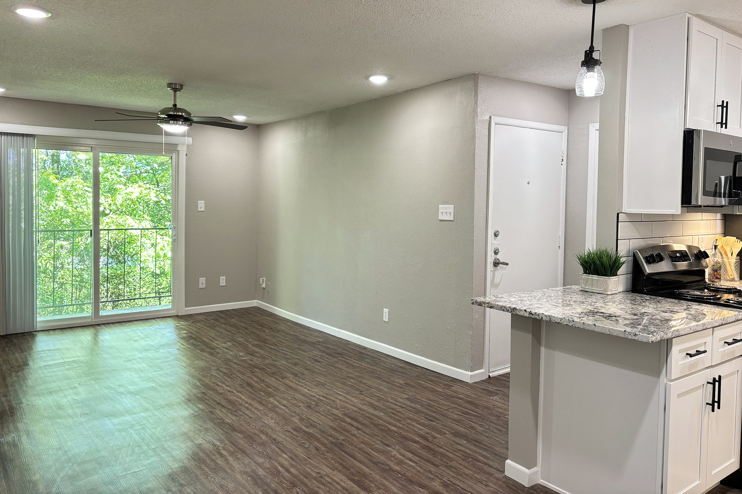 A spacious and bright living area featuring a ceiling fan, modern kitchen with granite countertops, and a sliding glass door that opens to a view of greenery outside. The walls are painted in soft gray, and the flooring is sleek and wooden, creating a welcoming atmosphere.