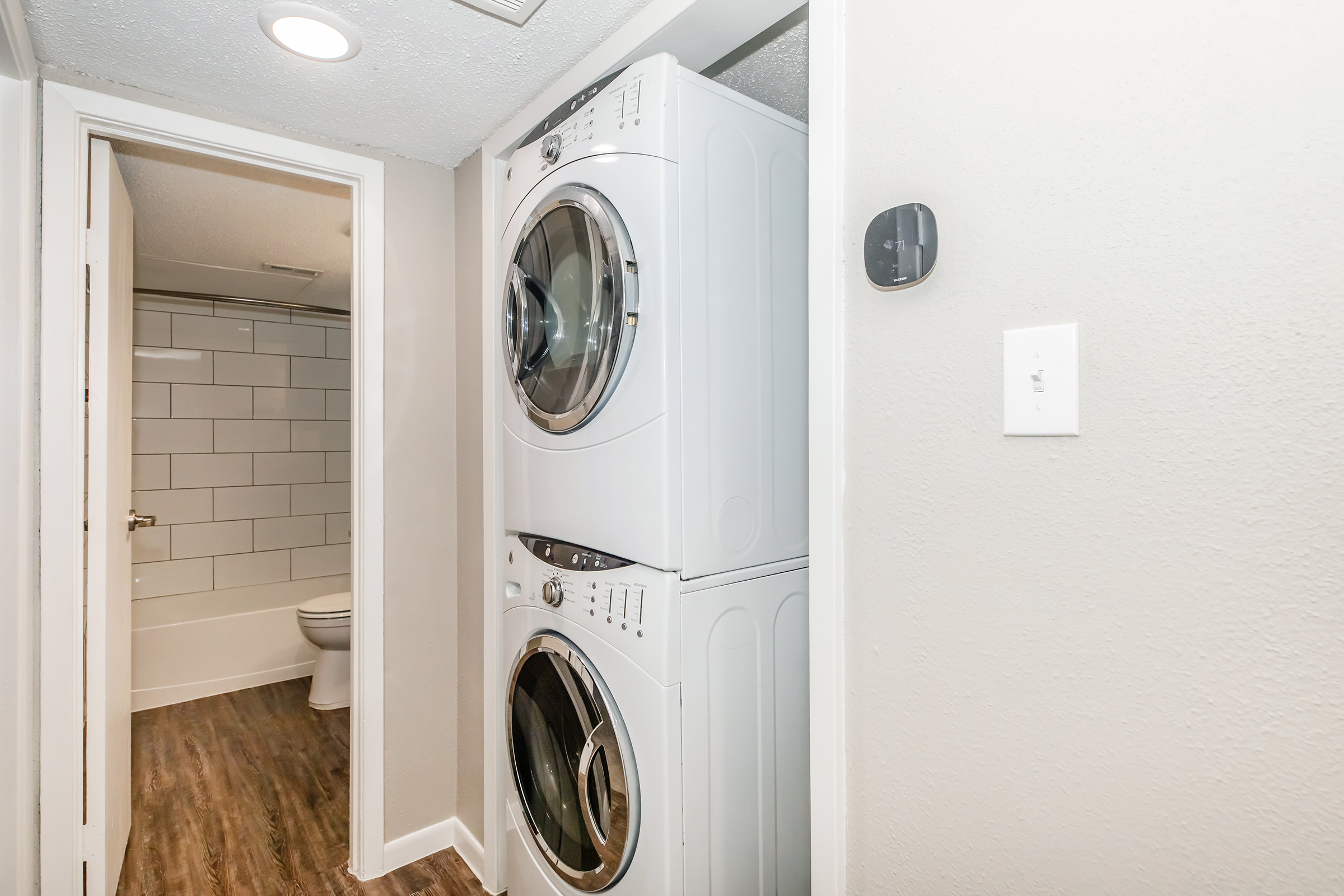 A laundry area featuring a stacked washer and dryer unit. The wall is painted a light color, and there is a partially visible bathroom with white tiled walls and a toilet in the background. The flooring is a wood-like laminate, giving the space a modern and clean appearance.
