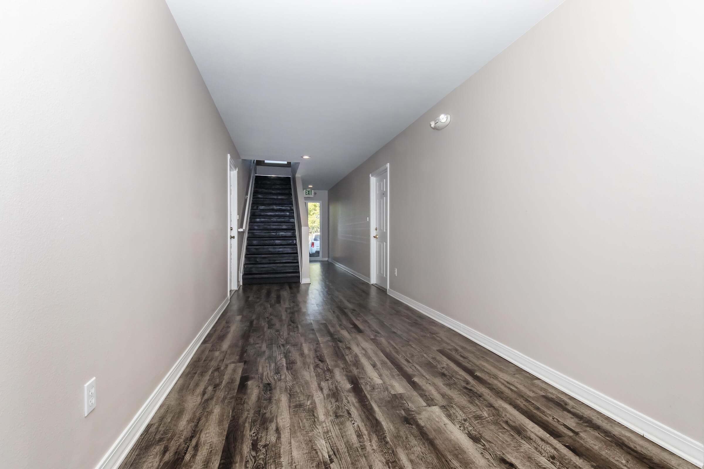A spacious, well-lit hallway featuring light gray walls and dark wood flooring. At the end of the hallway, a staircase with black steps leads to the upper level. Doorways are visible on the right side, and a window allows natural light to enter the space.