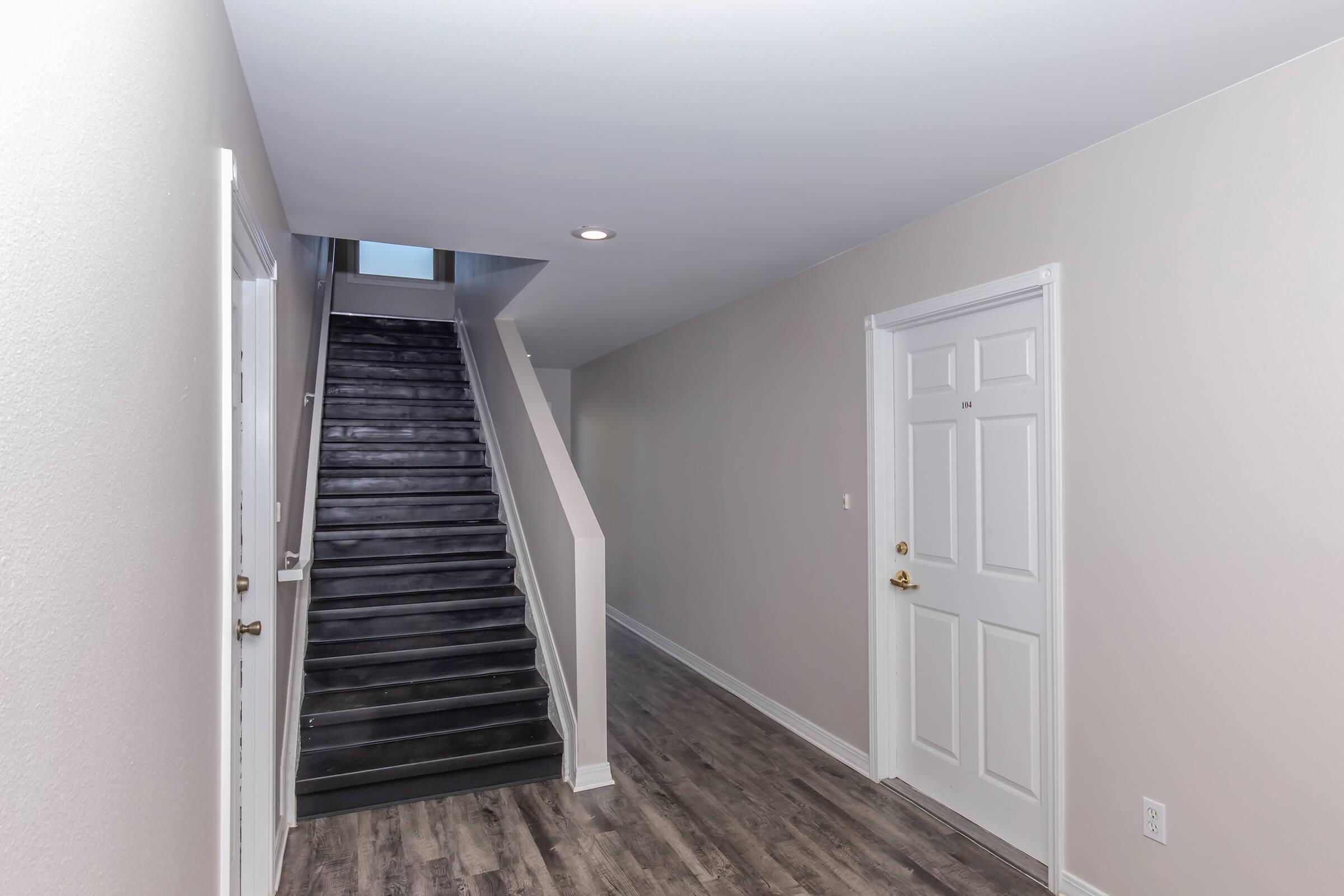 A bright hallway with light-colored walls and dark wooden flooring. A staircase with a contemporary design leads upward, illuminated by a window above. A white door is visible on the right side of the hallway, contributing to a clean and modern aesthetic.