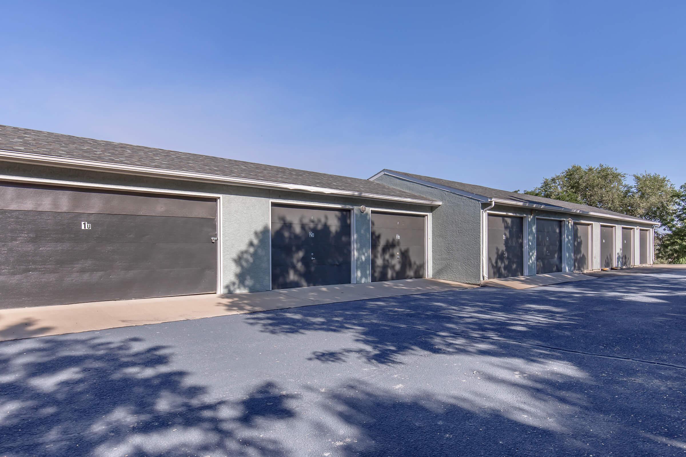 A row of garage doors in a residential area, with shadows cast on the driveway from nearby trees. The clear blue sky above adds to the bright and open atmosphere. Each garage is uniformly styled, showing a well-maintained property.