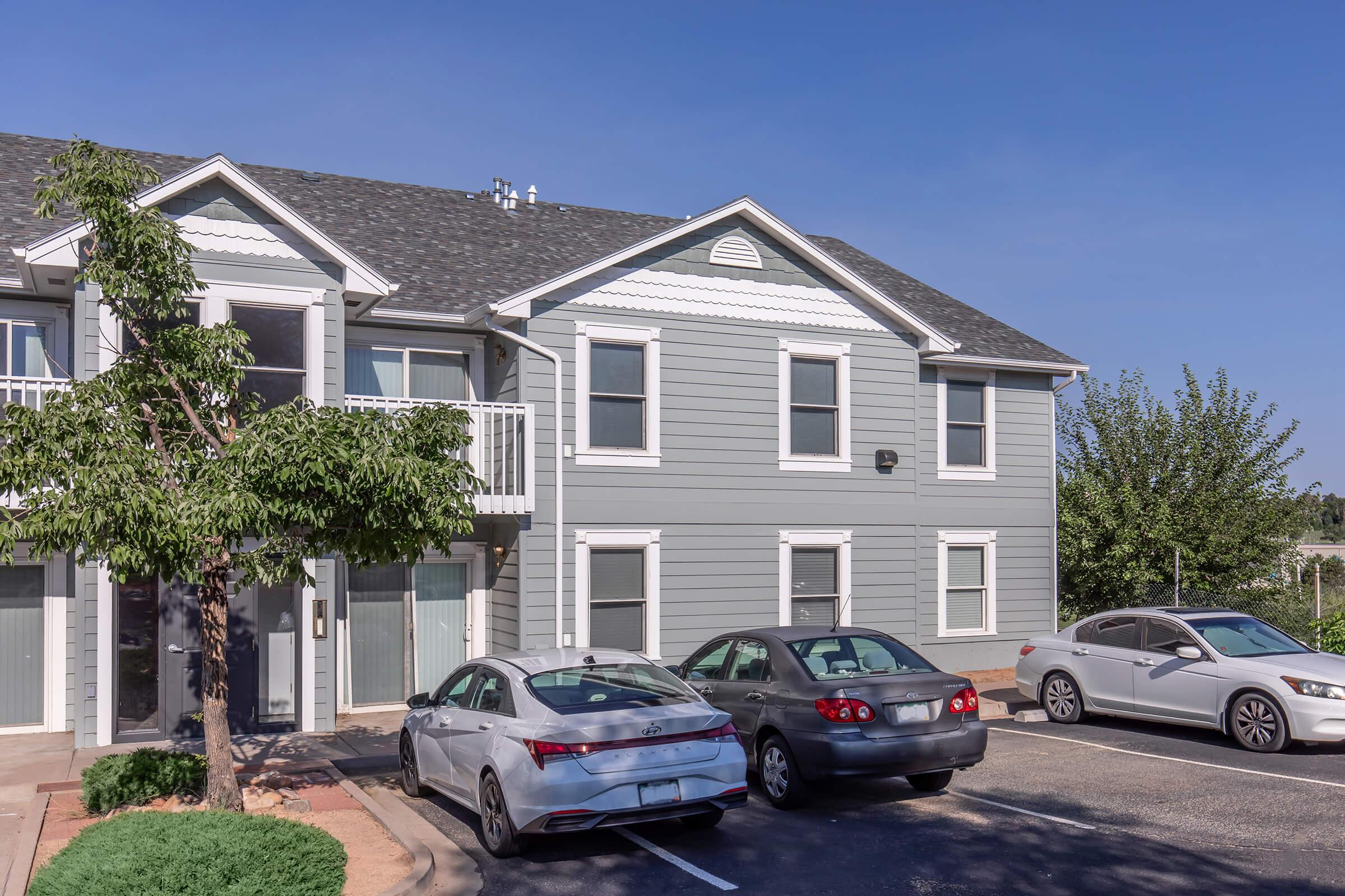 Gray two-story apartment building with white trim, featuring a balcony on the upper level. A tree is visible in the foreground, and three cars are parked in front of the building. The setting is sunny with a clear blue sky.
