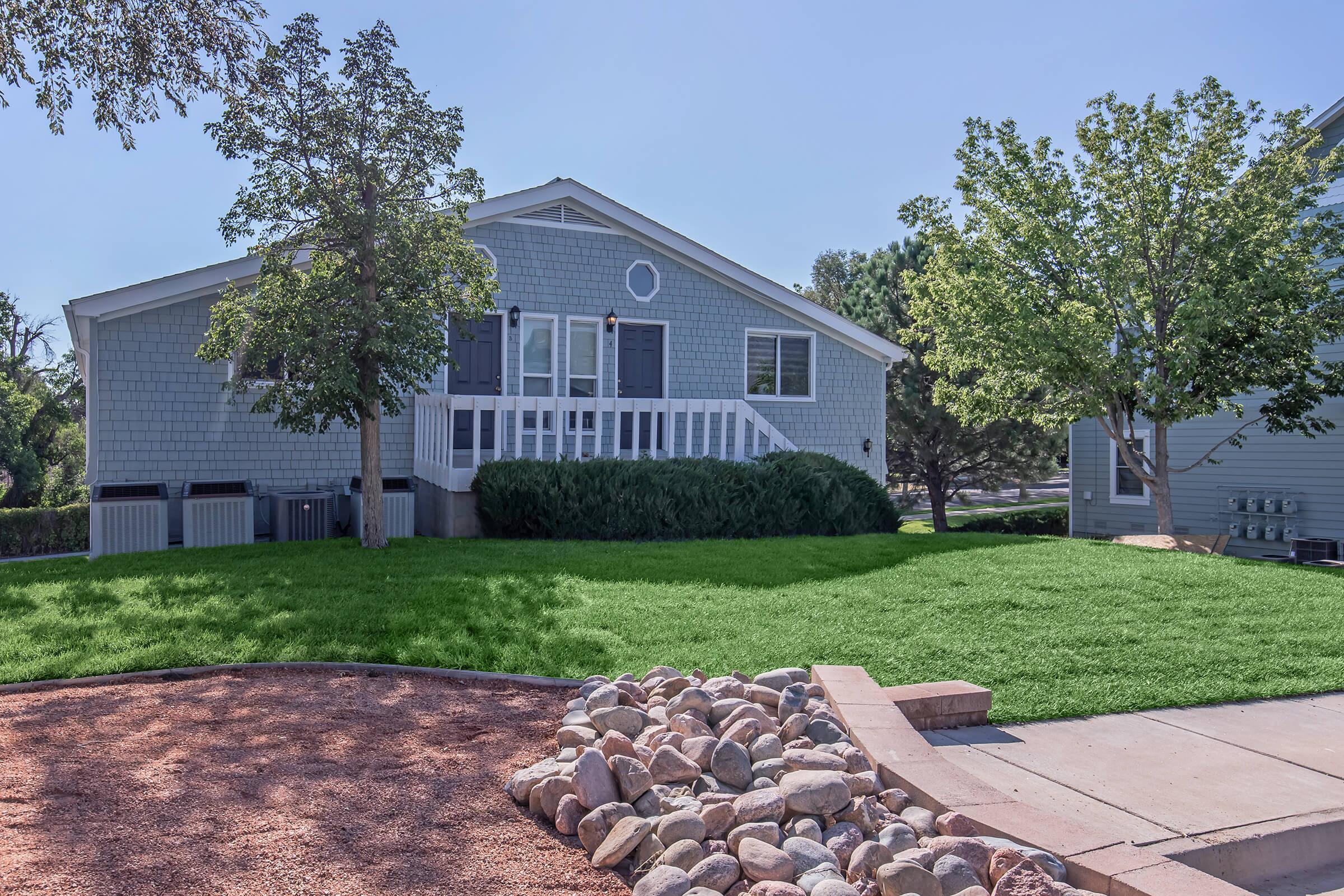 A light gray house with white trim and a front porch, surrounded by lush green grass and trees. Decorative rocks line the walkway, leading to the entrance. The house is set in a residential neighborhood, with other homes visible in the background, all under a clear blue sky.