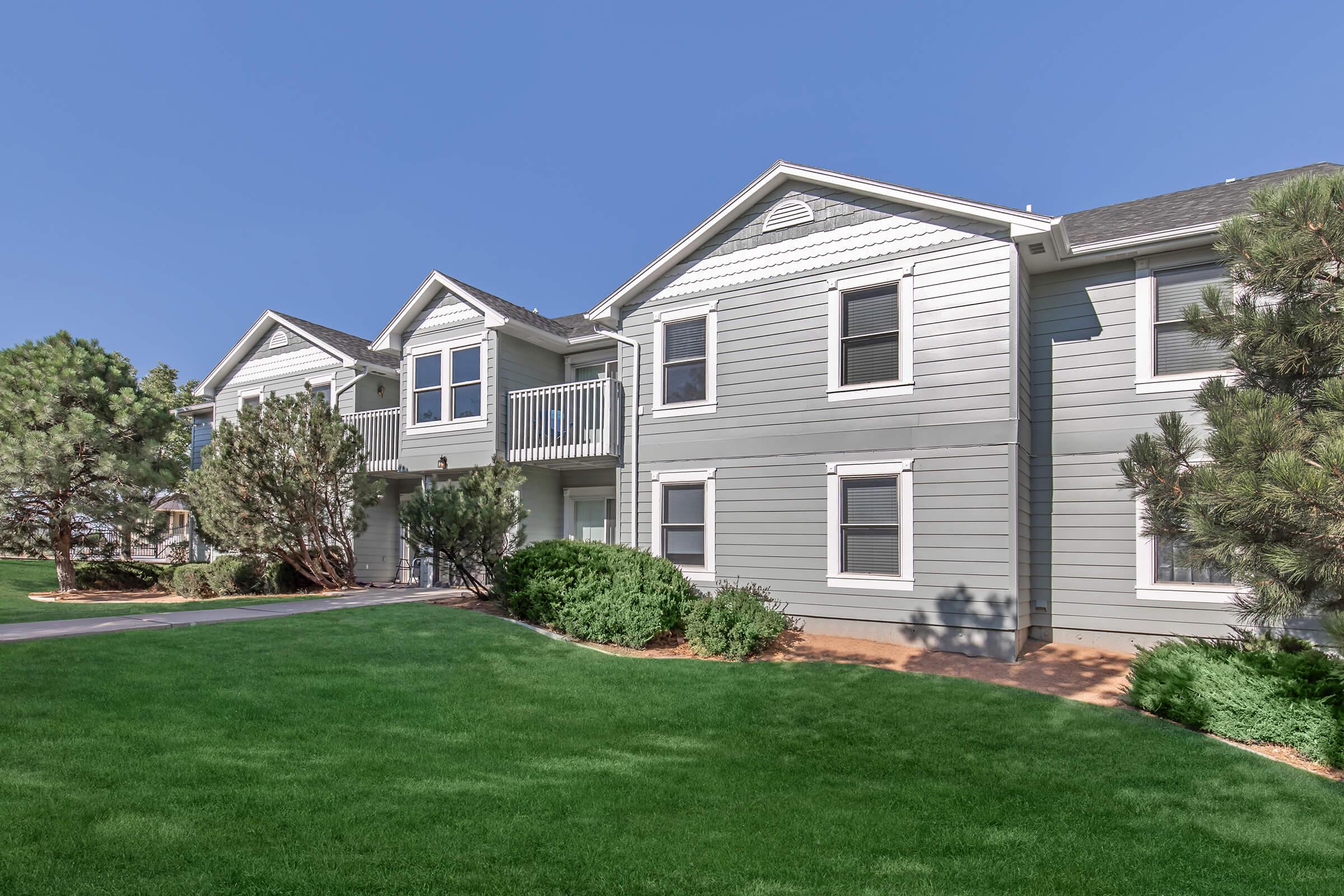 A scenic view of a modern, light gray apartment building with multiple units. The building features white trim and balconies, surrounded by well-maintained green grass and small trees. Clear blue sky above enhances the sunny atmosphere of the residential area.