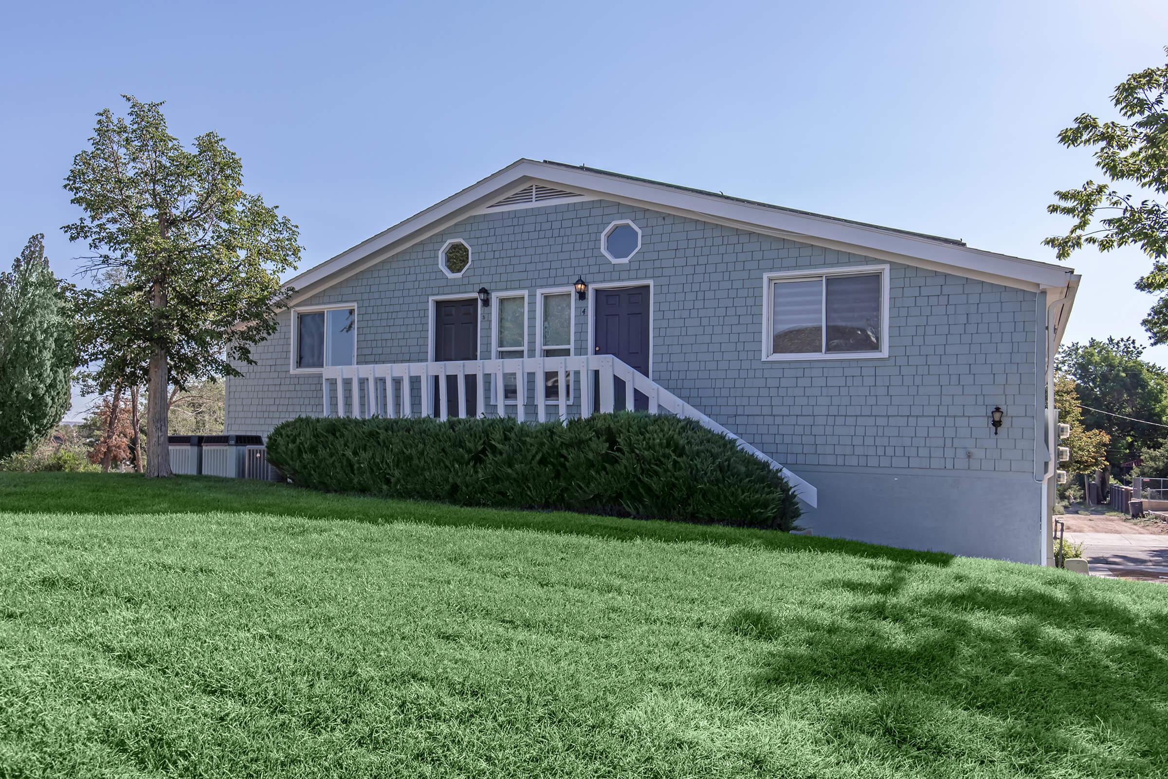 A light blue house with a gray front door and a white railing, situated on a grassy slope. The house has decorative circular windows and is surrounded by greenery, with trees on the left and a clear blue sky in the background. The scene captures a bright and inviting residential setting.