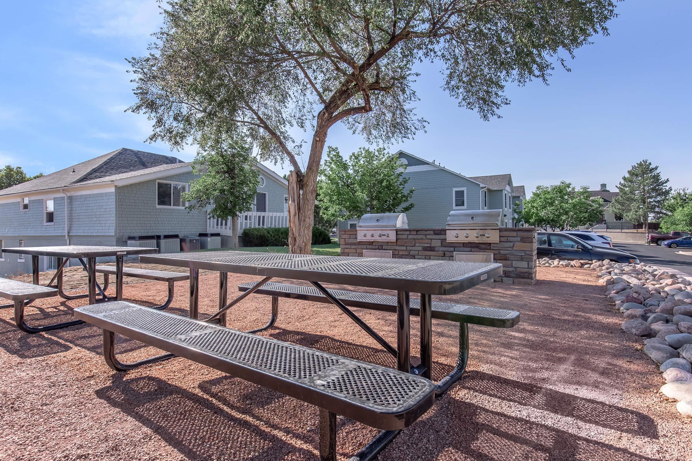 A picnic area featuring a large metal table and benches surrounded by gravel. In the background are two residential buildings and a green tree. There are two barbecue grills nearby, and additional parking spaces can be seen in the distance. The scene is well-lit by natural sunlight.