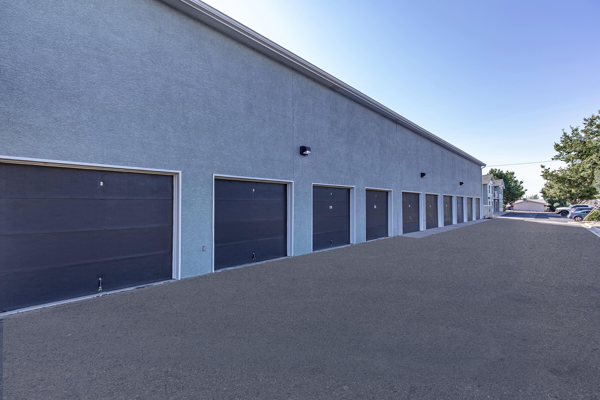 A row of closed garage doors on a gray building, set against a clear blue sky. The surface in front is paved, with no vehicles present, and there are trees visible in the background. The image conveys a sense of storage or commercial space.
