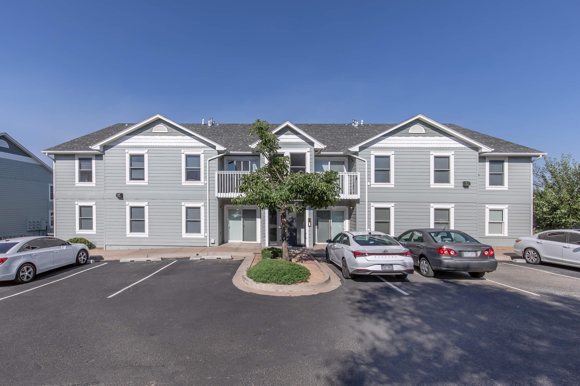A two-story apartment building with a light gray exterior. The front features balconies and several windows, with a small tree in the center of the entrance area. There are parked cars in the foreground and a clear blue sky above, creating a bright, inviting atmosphere.