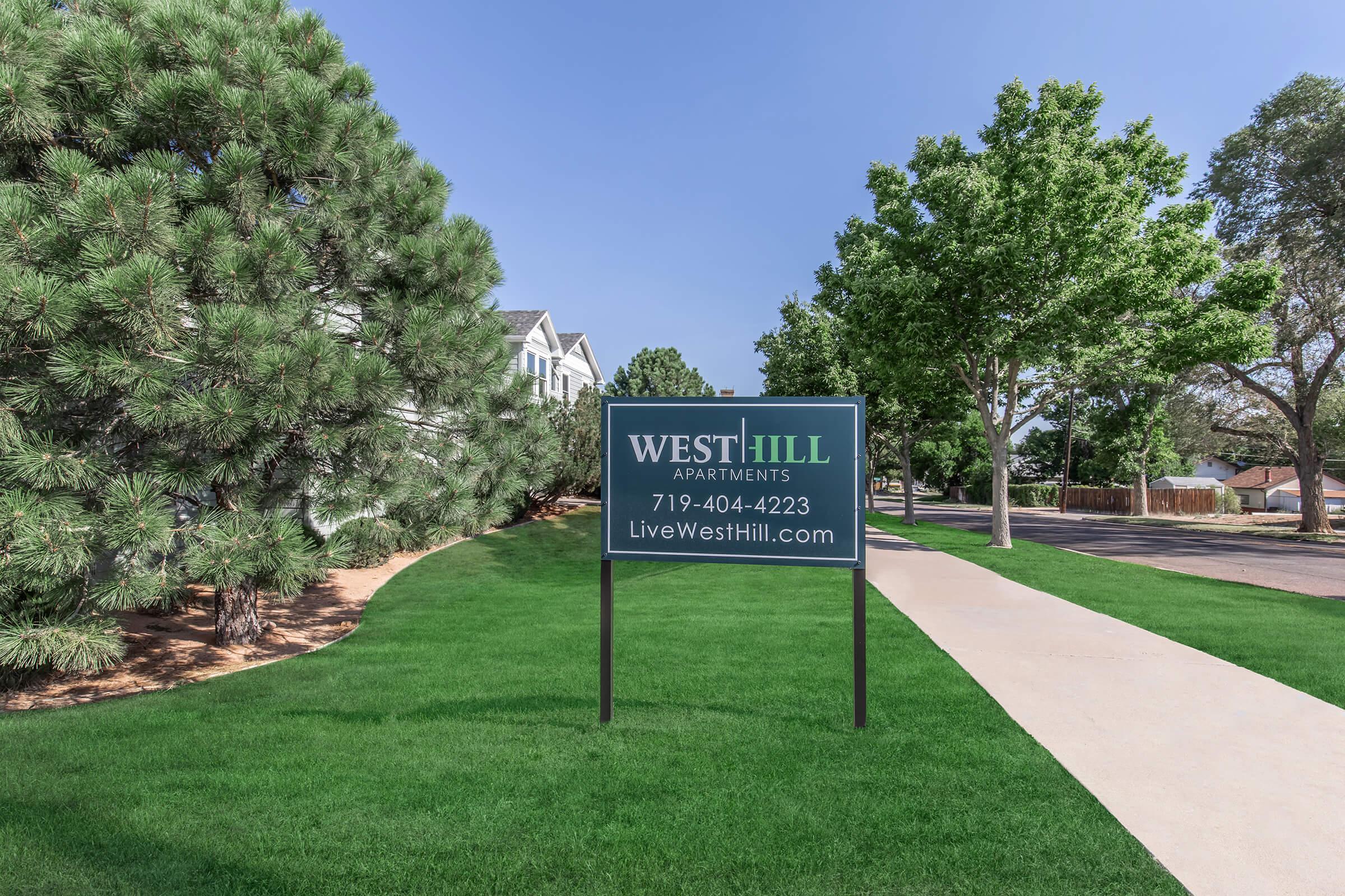 A sign for West Hill Apartments is displayed on a grassy area, featuring the contact number 719-404-4223 and the website LiveWestHill.com. In the background, there are trees and a pathway, suggesting a residential neighborhood setting.