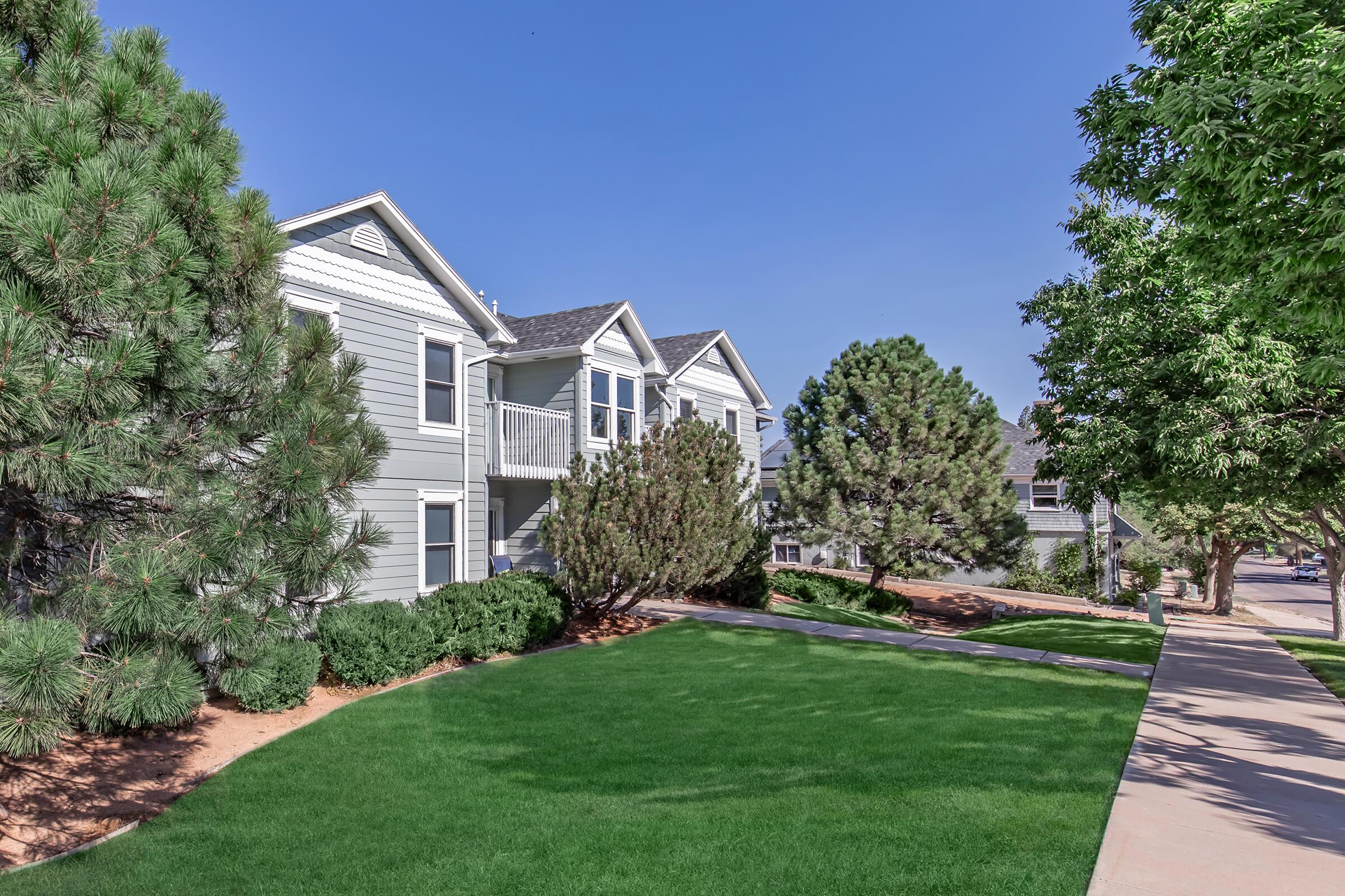 A residential area featuring two-story gray apartment buildings with green lawns, bordered by neatly trimmed bushes and trees. The sky is clear and blue, and a paved walkway runs alongside the buildings, creating a welcoming atmosphere.