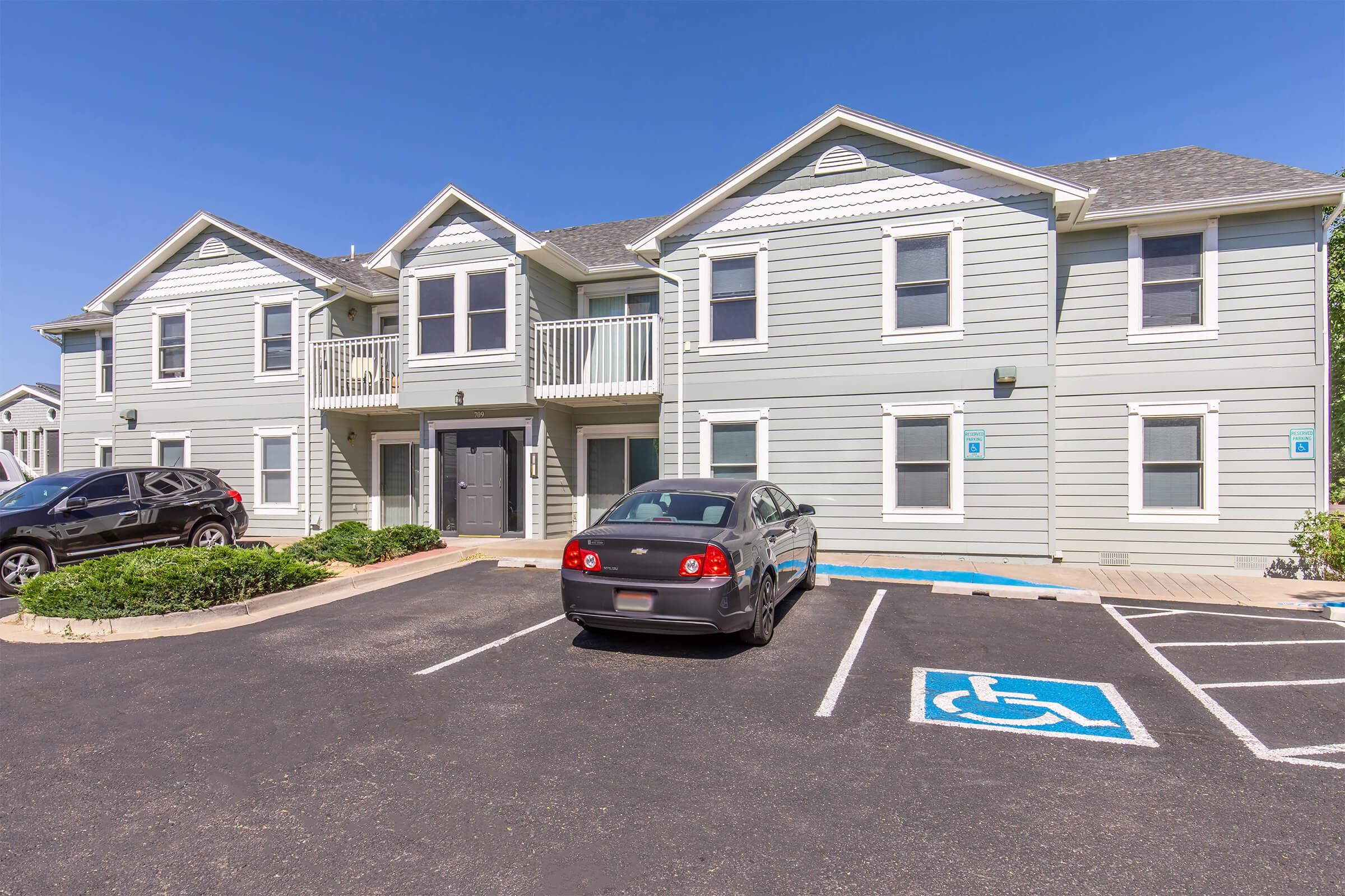 A two-story apartment building with light green siding. The front features a set of stairs leading to upper units, a couple of parked cars, and a designated handicapped parking space. The sky is clear and blue, creating a bright and inviting atmosphere.