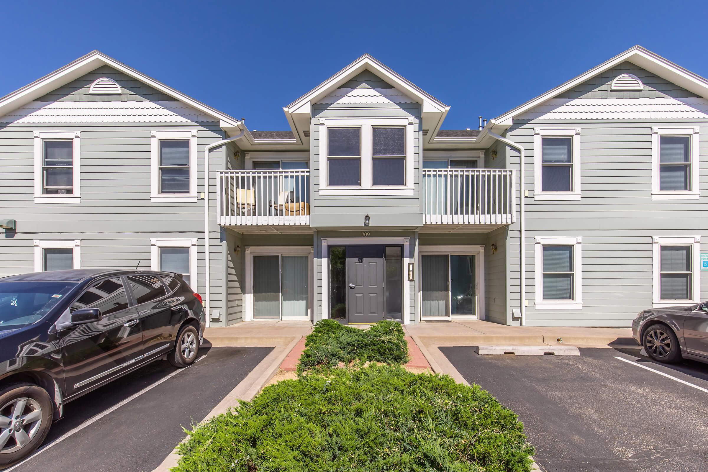 Exterior view of a two-story apartment building featuring gray siding and decorative white trim. The center unit has a double window and a balcony. Two parking spaces are visible in front of the building, alongside a neatly trimmed green shrub. Clear blue sky above.