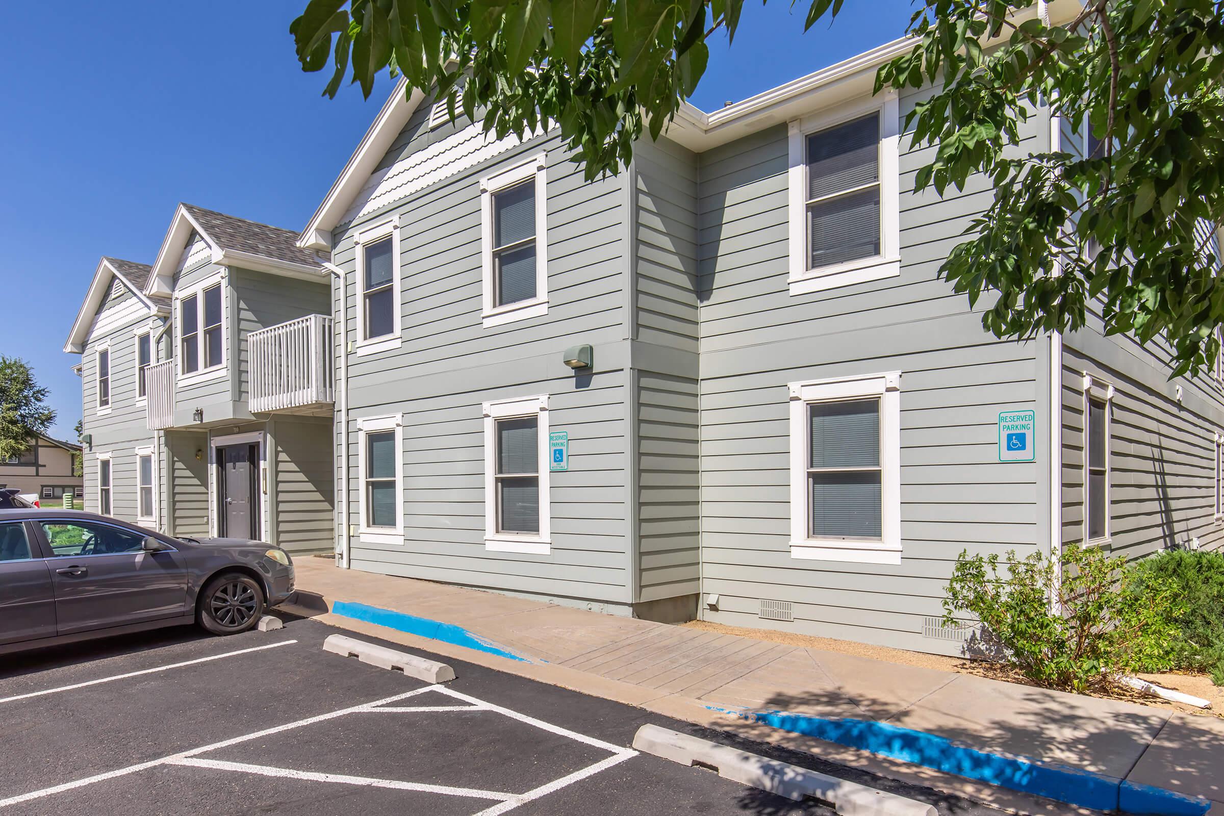 Two-story light gray residential building with white trim, featuring several windows and a balcony on the left side. A gray car is parked in front, and there are blue parking spaces marked on the asphalt. Green foliage is visible in the foreground, under a clear blue sky.