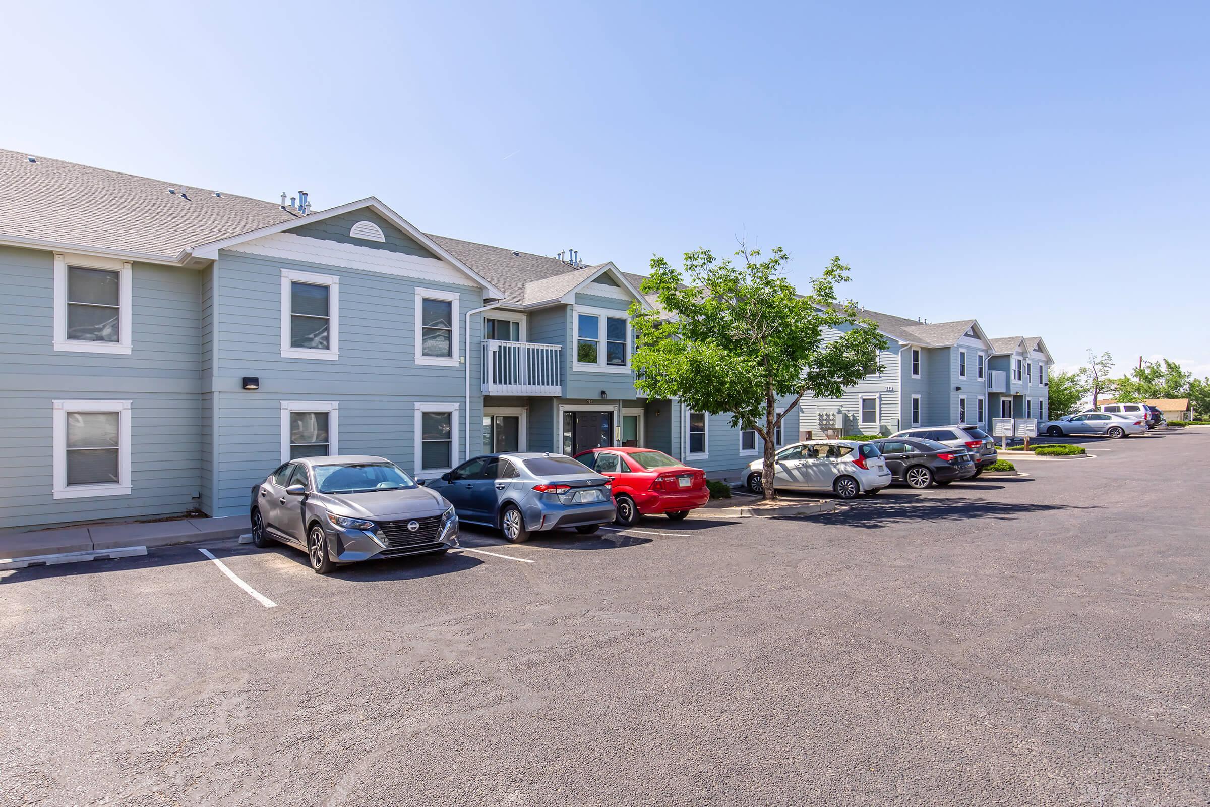 Row of multi-unit residential buildings with light blue siding and parked cars in the foreground. The parking lot is full with various vehicles, and there are a few trees providing shade. The sky is clear and blue, indicating a sunny day.