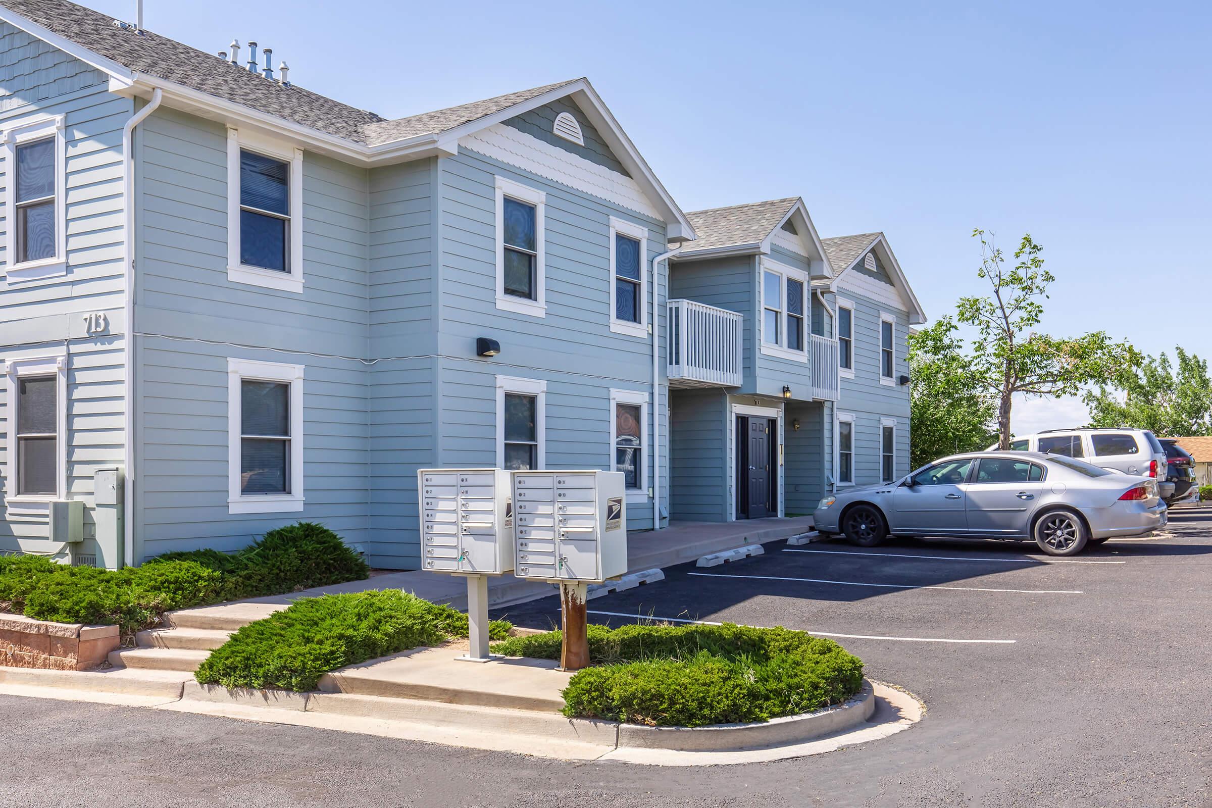 A light blue two-story apartment building with several windows and a balcony. In front, there are organized mailboxes and a parking lot with cars. Green shrubs line the walkway, and the sky is clear with few clouds. The overall setting appears well-maintained and inviting.