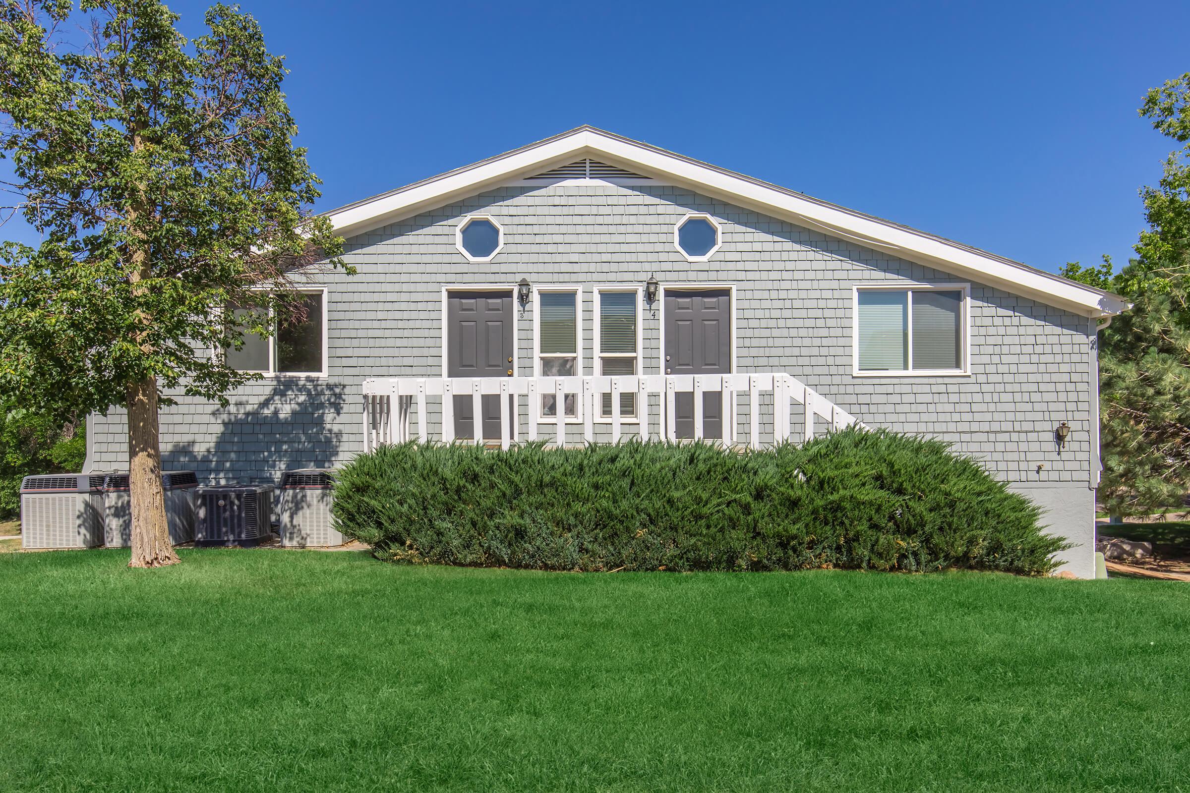 A gray house with a sloped roof, featuring two front doors and circular windows. The house is surrounded by a well-maintained green lawn and a row of shrubs in front. A small air conditioning unit is visible to the left of the entrance. The sky is clear and blue, suggesting a sunny day.
