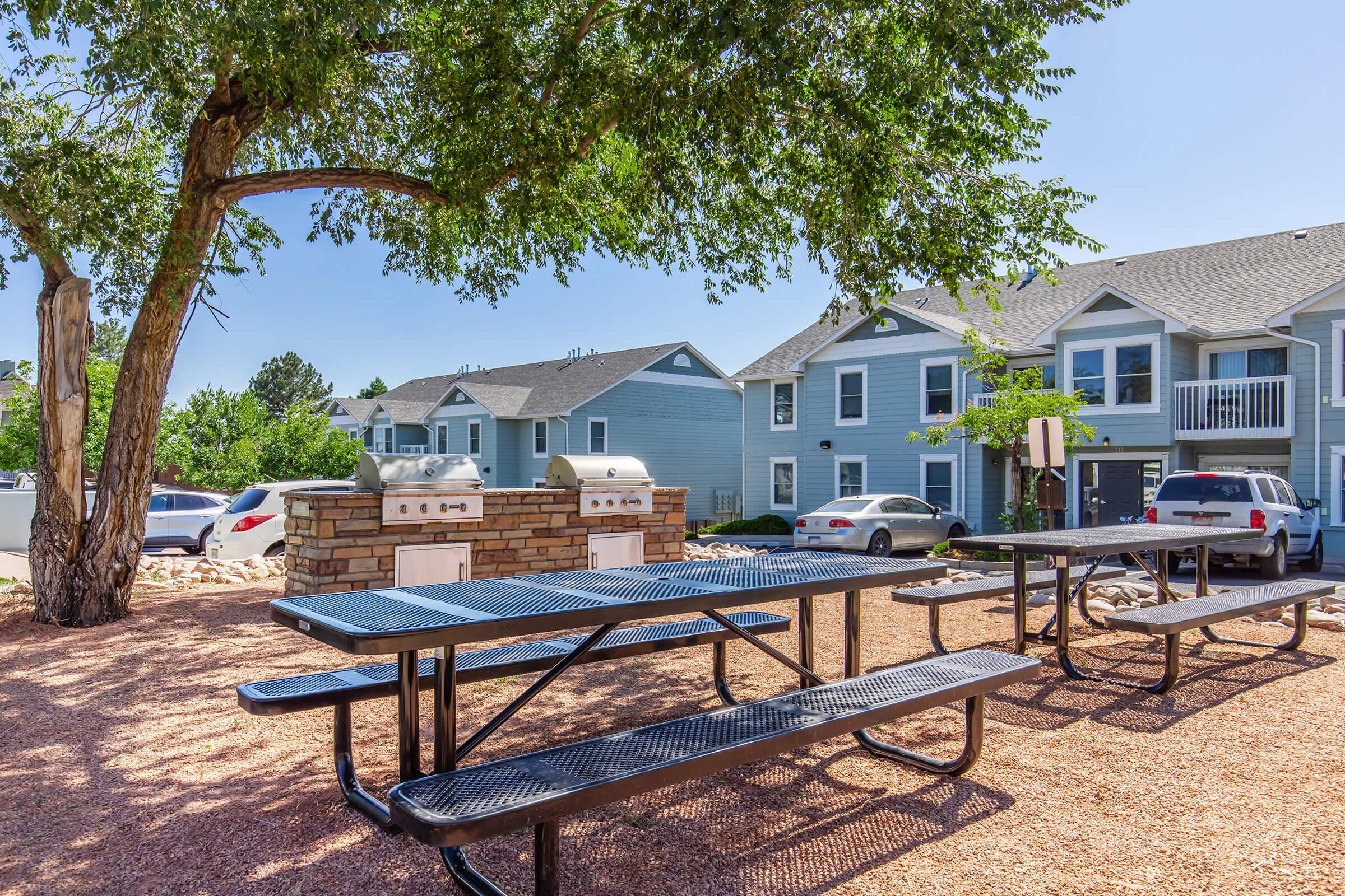 A picnic area featuring black metal tables and grills under a large tree. In the background, there are two-story residential buildings painted in light blue. Several parked cars are visible nearby, and the ground is covered with gravel. The scene is set on a sunny day.